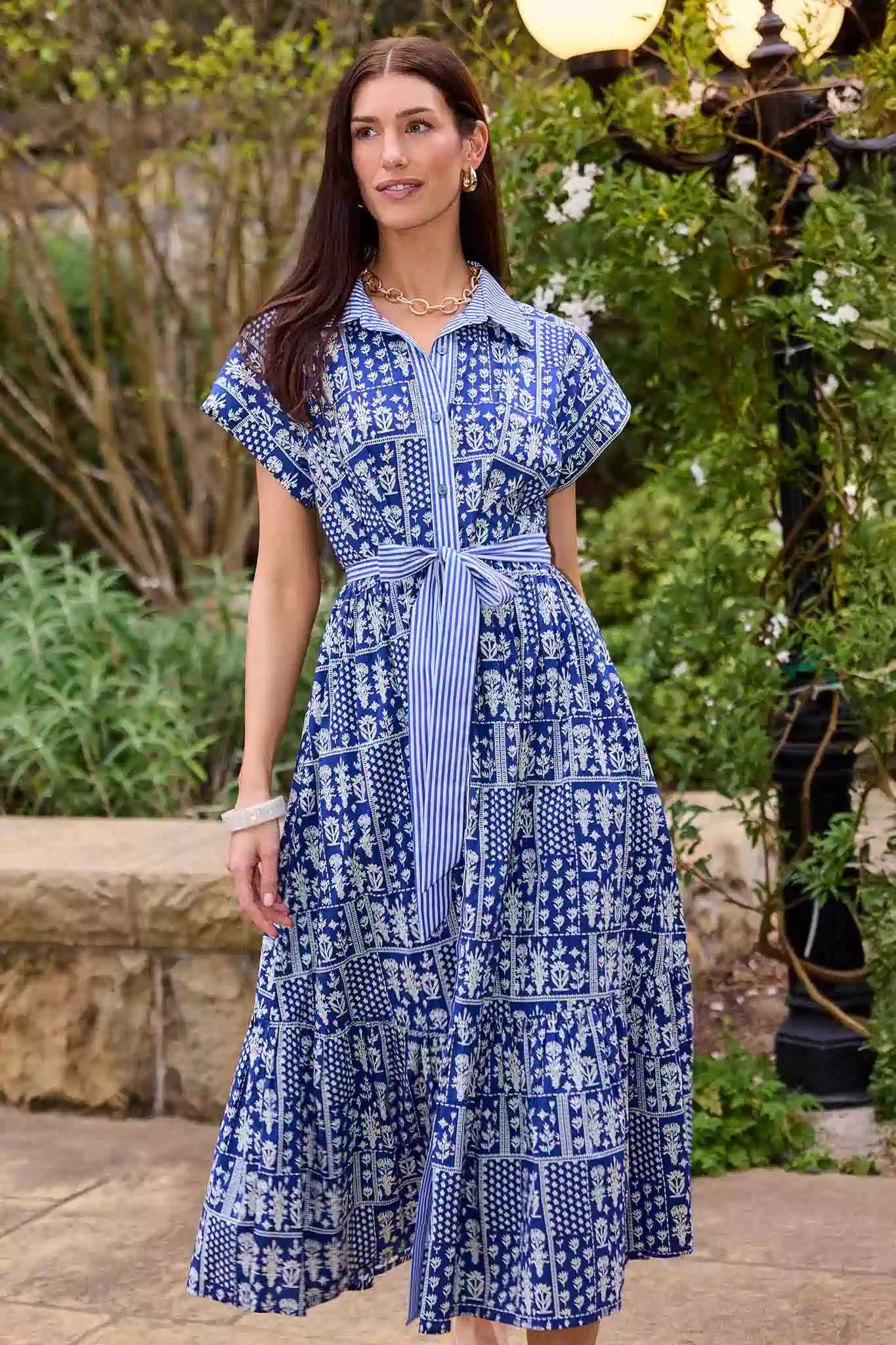 Woman wearing a blue patterned dress standing outdoors with greenery in the background