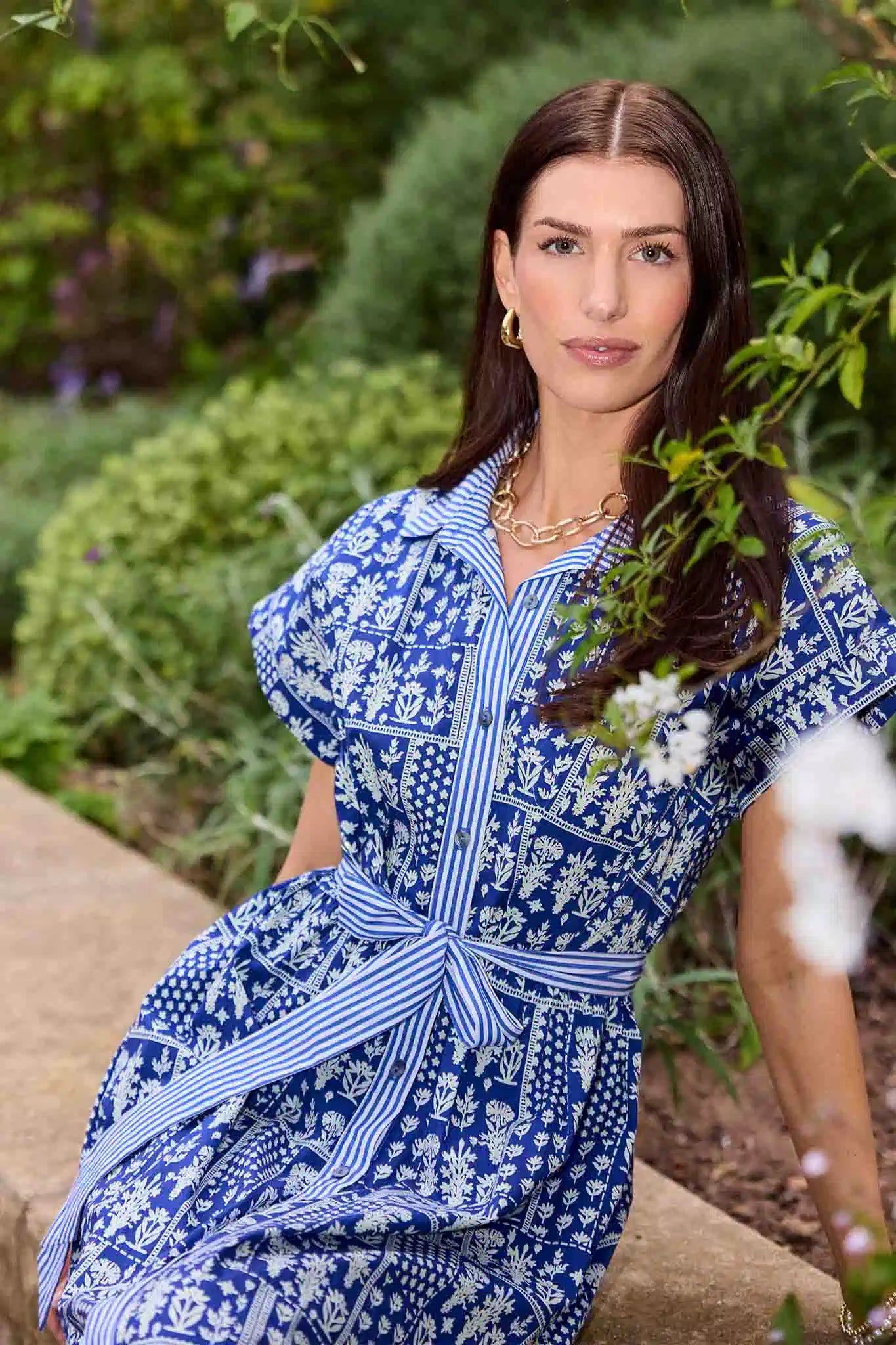 Woman wearing a blue and white patterned dress sitting outdoors with greenery in the background
