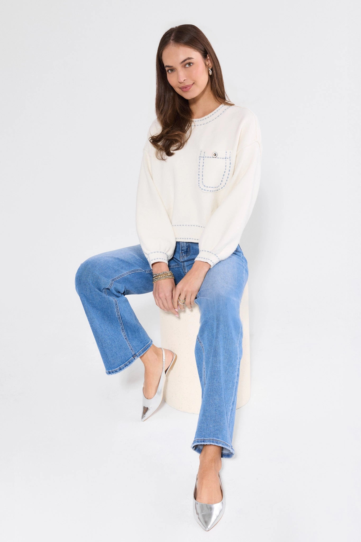 Woman wearing a white sweatshirt and blue jeans sitting on a white surface with a white background