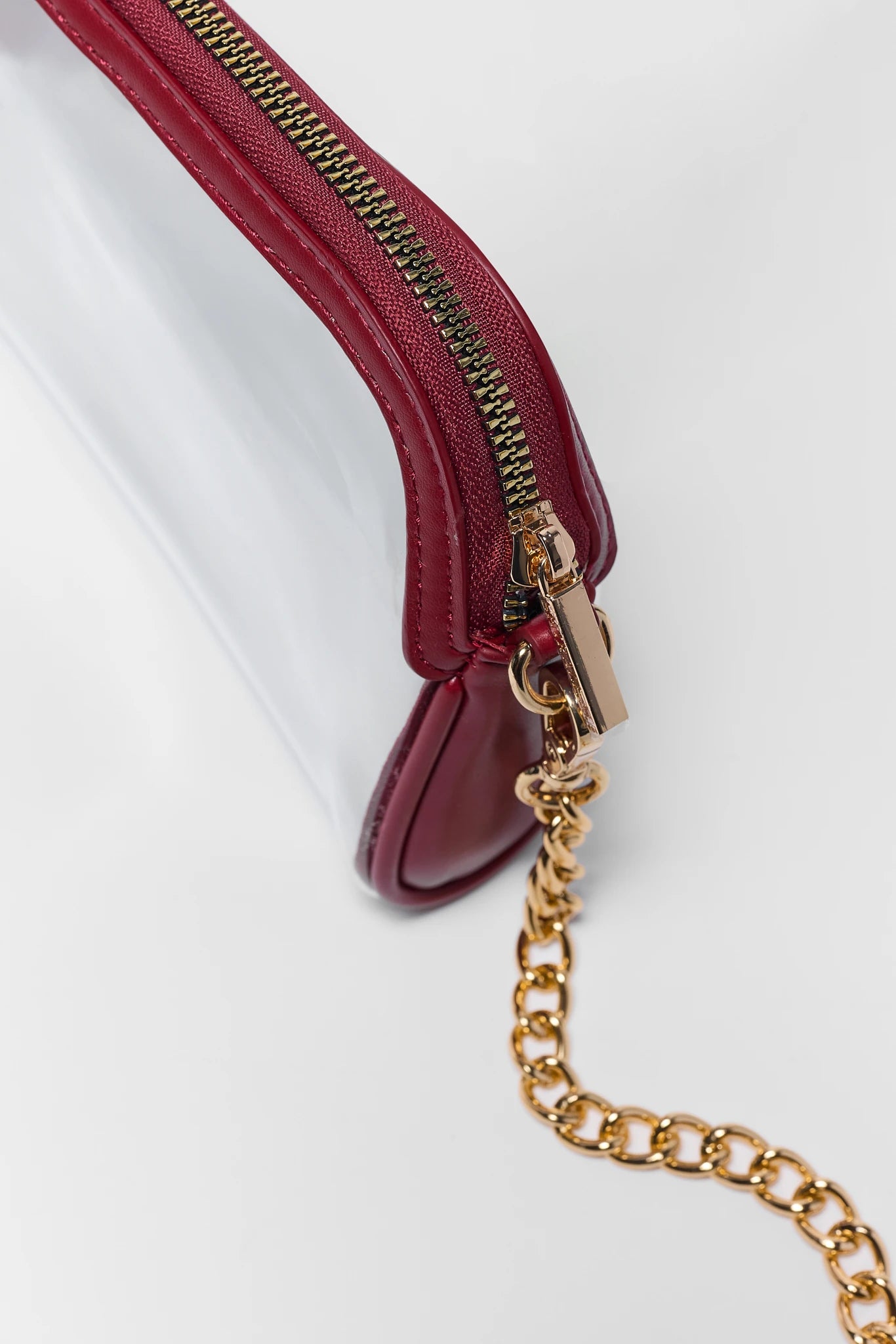 Close-up of a burgundy leather zipper pouch with a transparent plastic body and a gold-tone chain strap, shown on a white background.