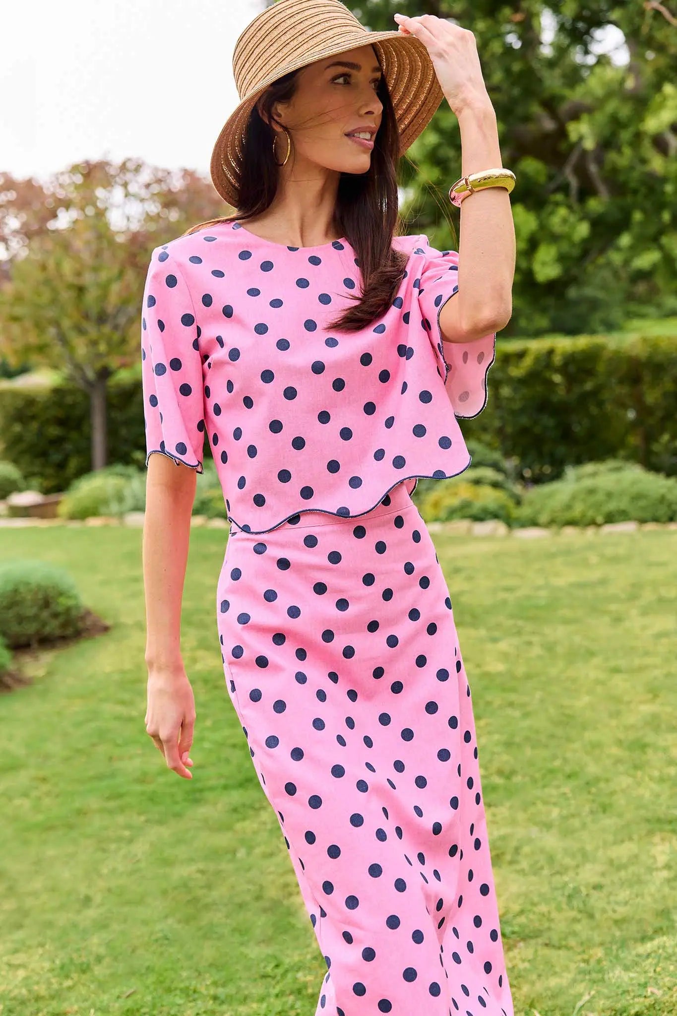 Woman in a pink polka dot outfit with a straw hat standing outdoors.