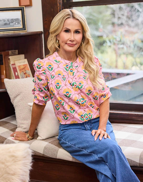 Woman sitting on a couch wearing a pink floral blouse and blue jeans, with a window and books in the background.