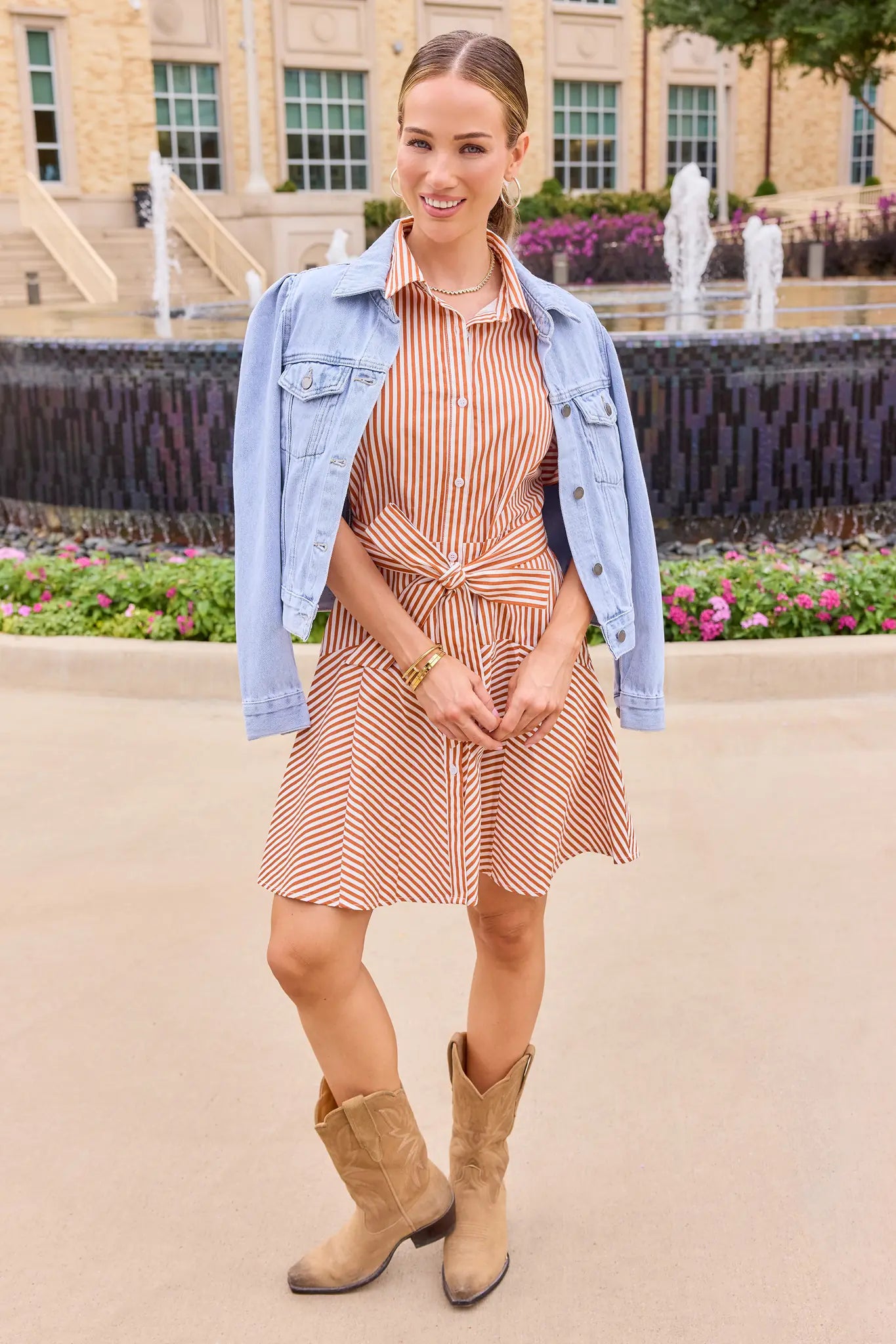 A woman stands outdoors wearing a light blue denim jacket over a rust-orange and white striped shirt dress with a tie waist, paired with tan cowboy boots. The background shows a fountain, flowers, and a stone building.