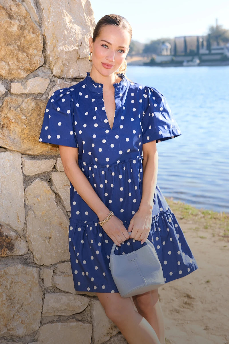 Woman in a blue polka dot dress standing by a stone wall with water in the background