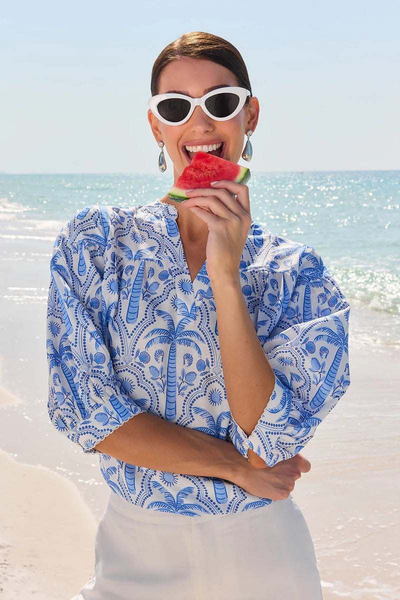Woman in a blue and white patterned top eating watermelon on a beach. mobile