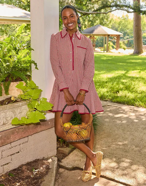 A woman stands outdoors on a walkway beside a white pillar, wearing a red and white gingham long-sleeve collared dress with red trim and holding a woven basket of fruit. The background shows a green lawn, trees, and a gazebo.