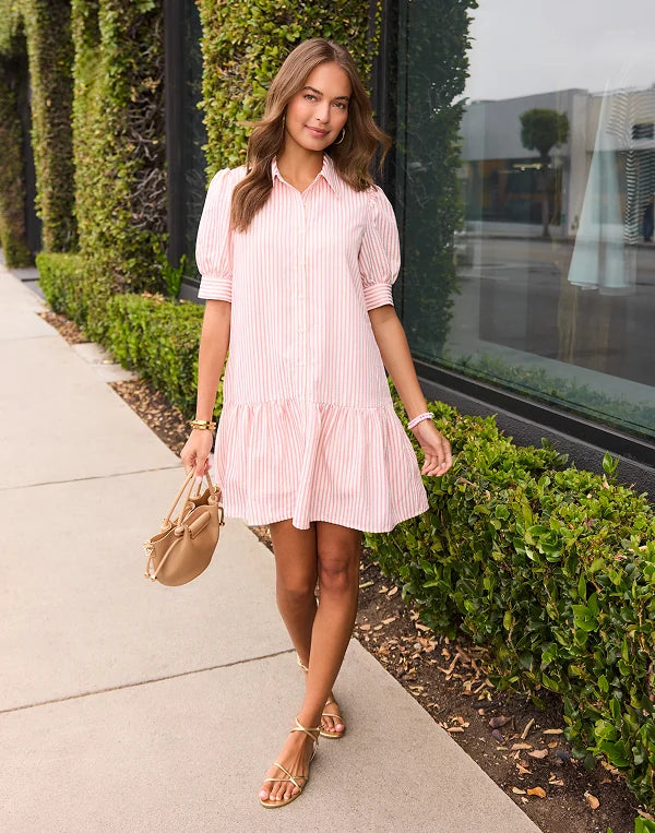 Woman in a pink dress standing on a sidewalk with greenery and a building in the background navigation