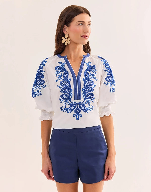 Woman wearing a white blouse with blue embroidery and blue shorts on a beige background navigation
