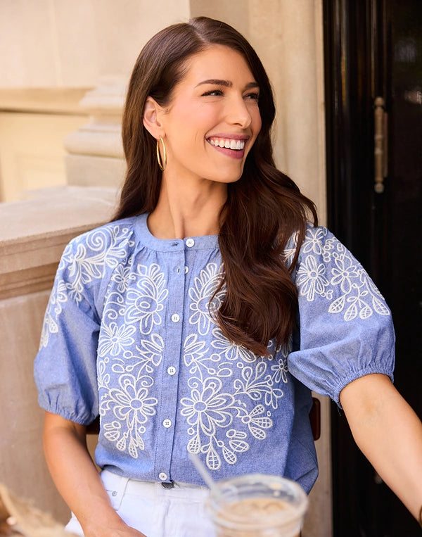 A woman smiling and looking to the side, wearing a light blue short-sleeve blouse with white floral embroidery and puff sleeves, paired with white pants. navigation