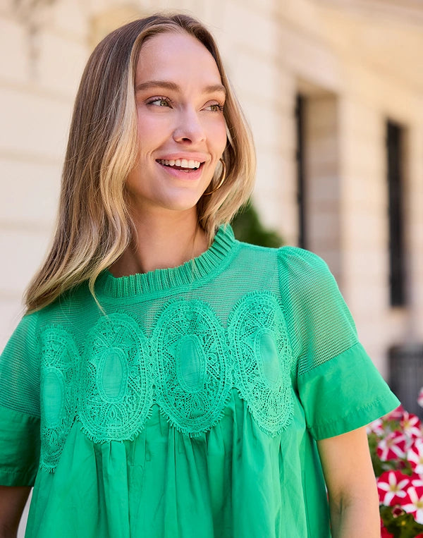 A woman wearing a bright green short-sleeve blouse with sheer mesh and lace detailing across the chest, photographed outdoors against a softly blurred building background. navigation