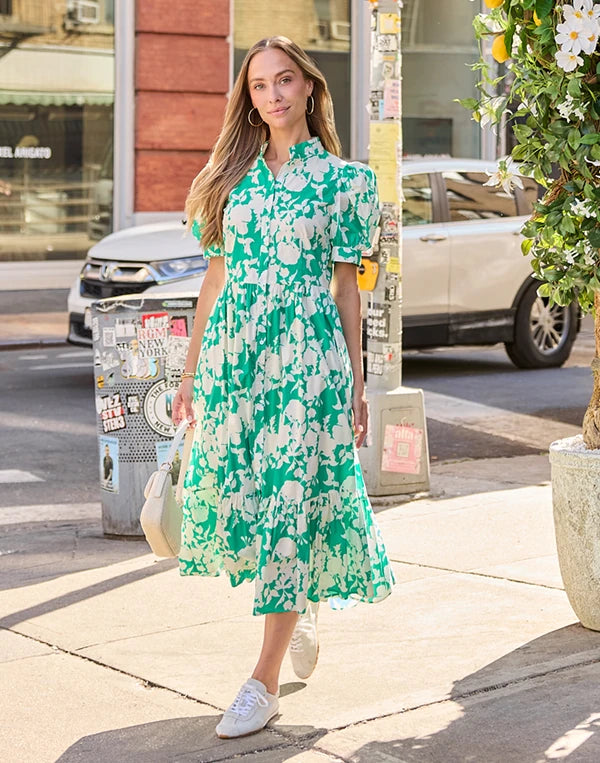 A woman walking on a city sidewalk wearing a green and white floral midi dress with short sleeves, a collared neckline, and a tiered skirt. She is carrying a light-colored handbag and wearing white sneakers. navigation