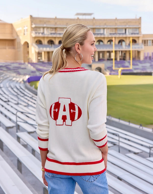 A woman stands in a stadium wearing a cream knit sweater with red and white striped trim and a large red 'A' graphic on the back, paired with blue jeans. navigation