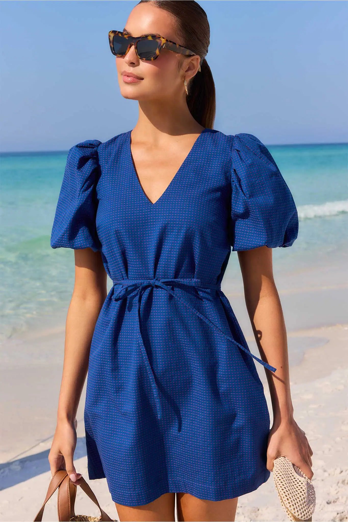 Woman in a blue dress standing on a beach with clear blue water and sky.