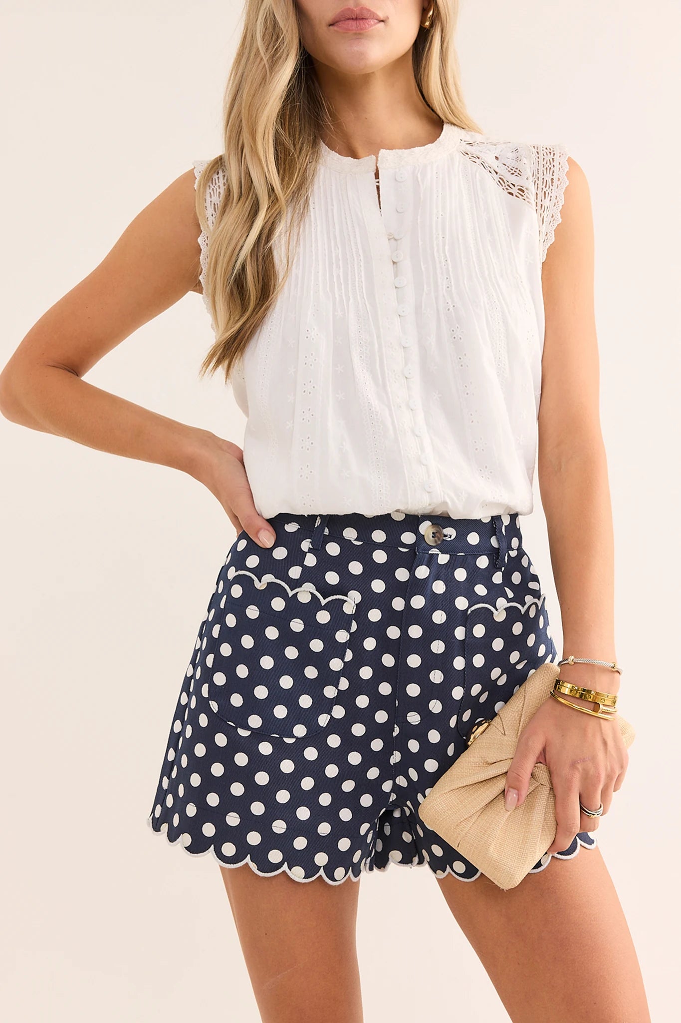 A woman modeling a sleeveless white top with lace shoulder details and navy blue shorts with white polka dots and scalloped hems, holding a beige clutch against a light beige background.