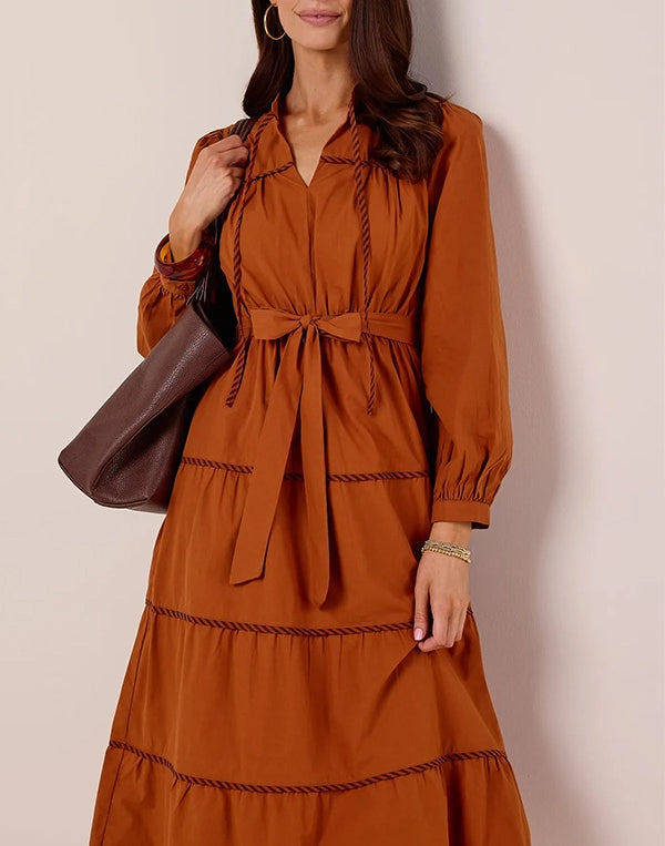 Woman wearing a rust-colored dress with a belt, holding a brown bag against a beige background