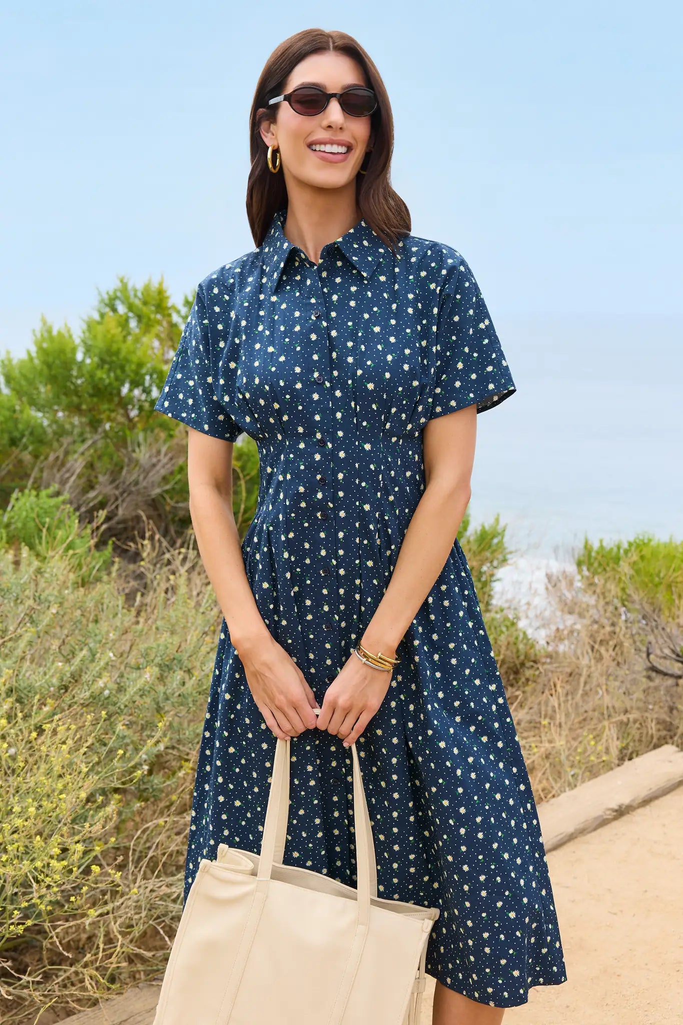 Woman wearing a navy dress with white polka dots, standing outdoors with greenery in the background.