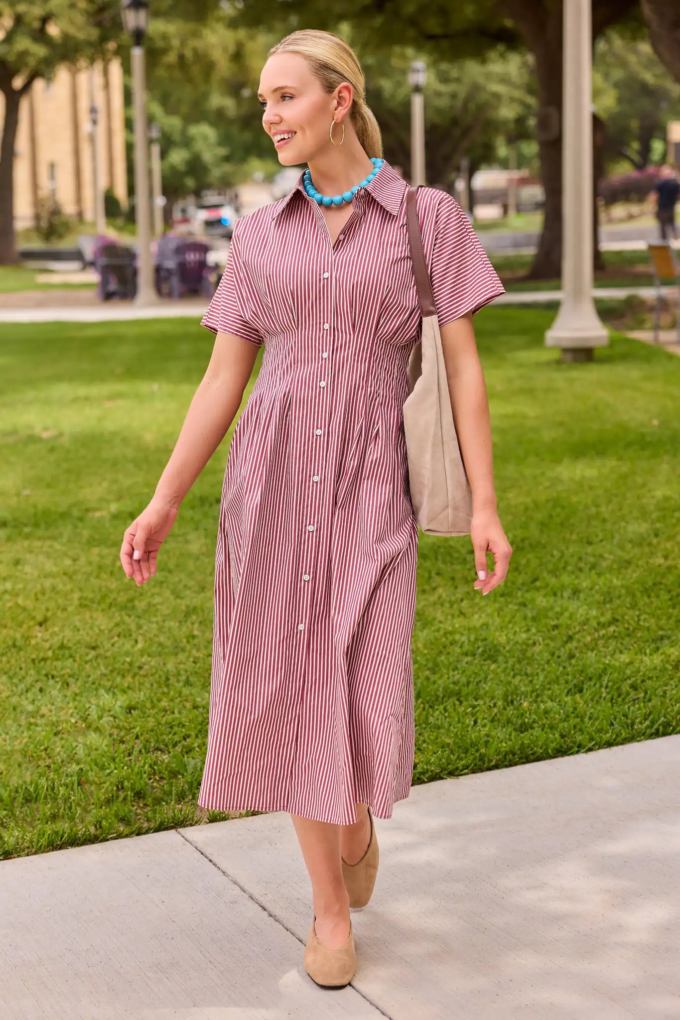 A woman walking outdoors on a sidewalk wearing a short-sleeve button-front midi dress with thin vertical pink and white stripes, a collared neckline, and a gathered waist. She is carrying a beige shoulder bag and wearing a turquoise necklace.