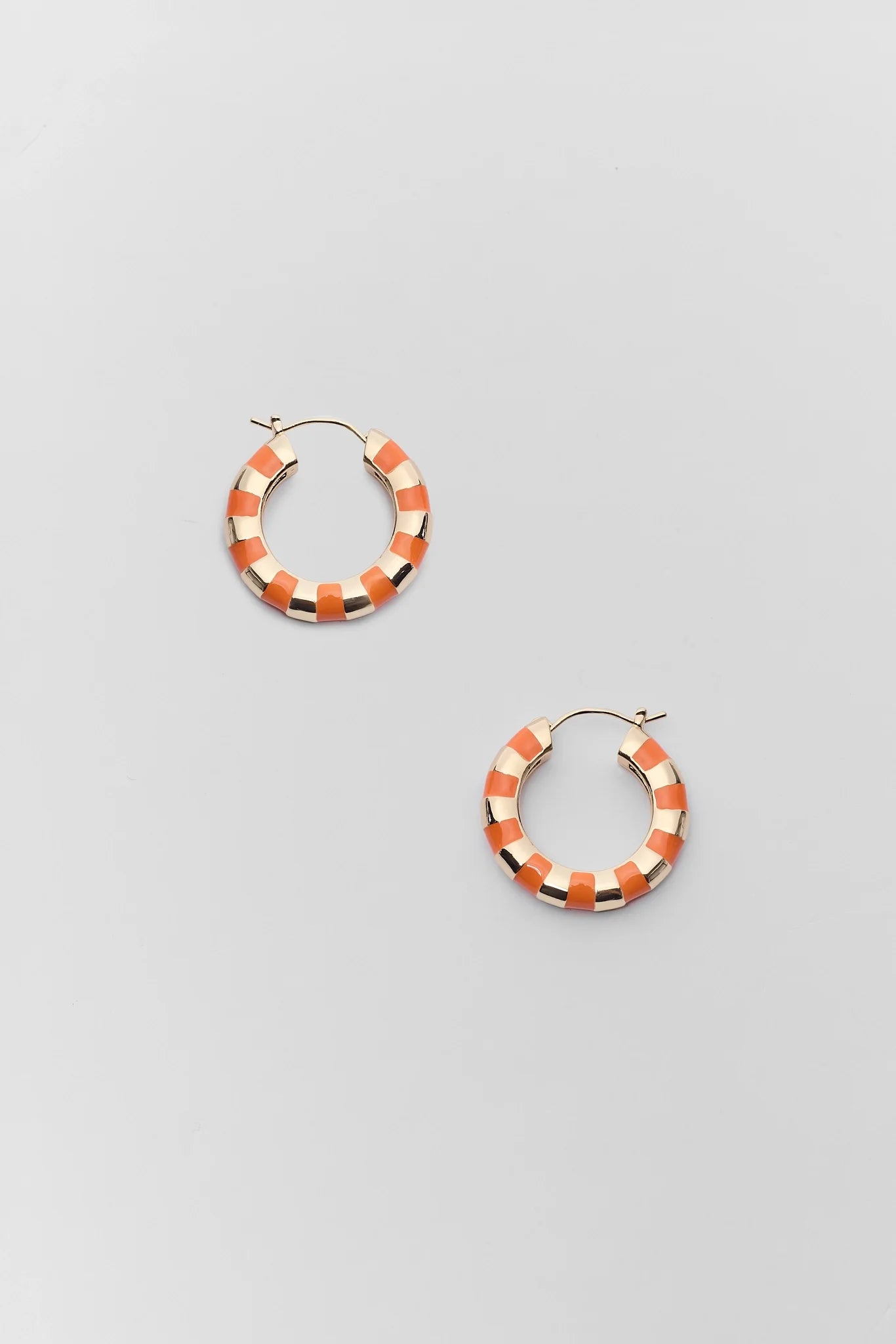A pair of gold hoop earrings with alternating orange and white enamel segments, displayed on a light gray background.