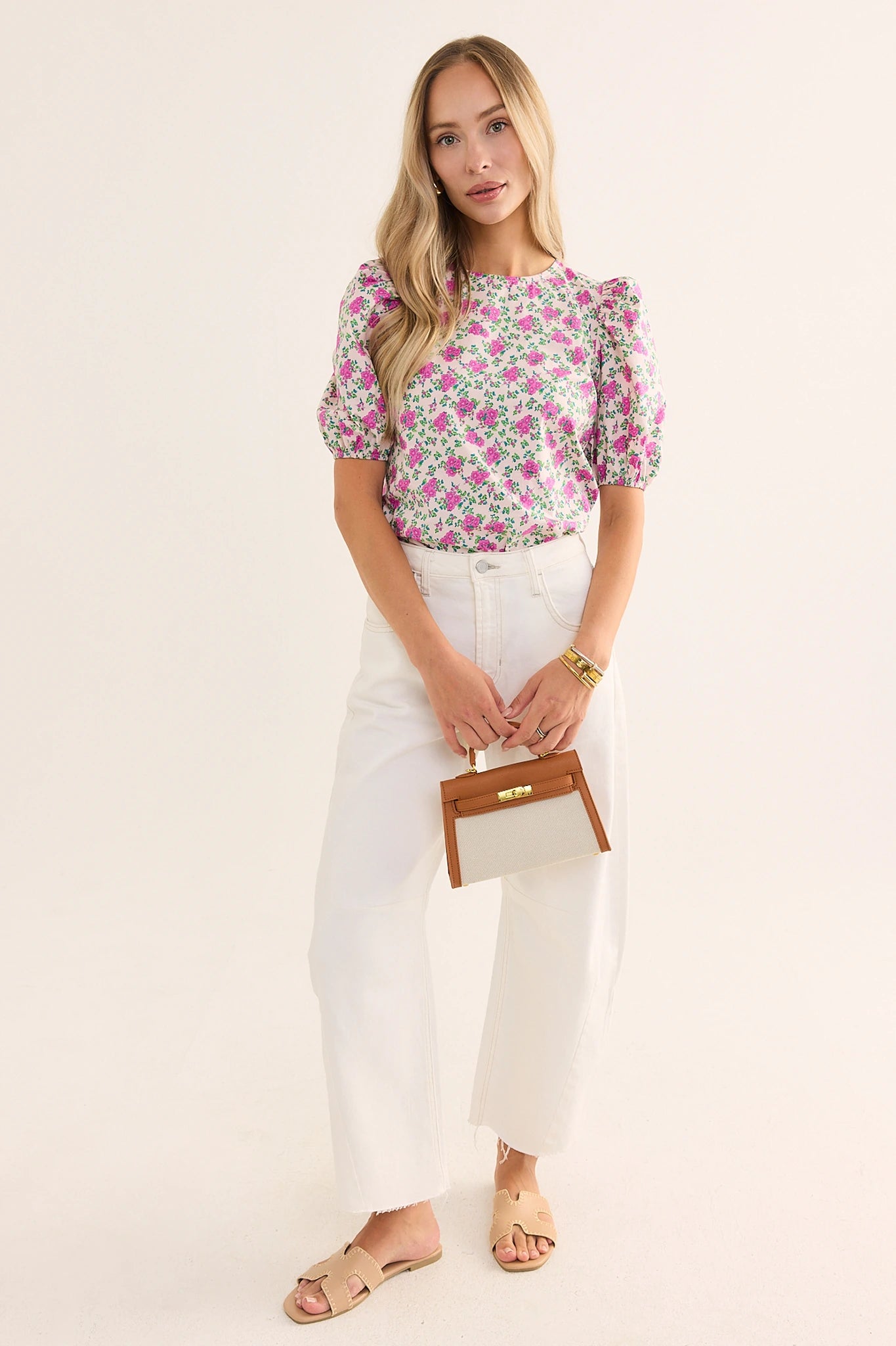 Woman wearing a floral blouse and white pants, holding a brown clutch against a plain background lifestyle