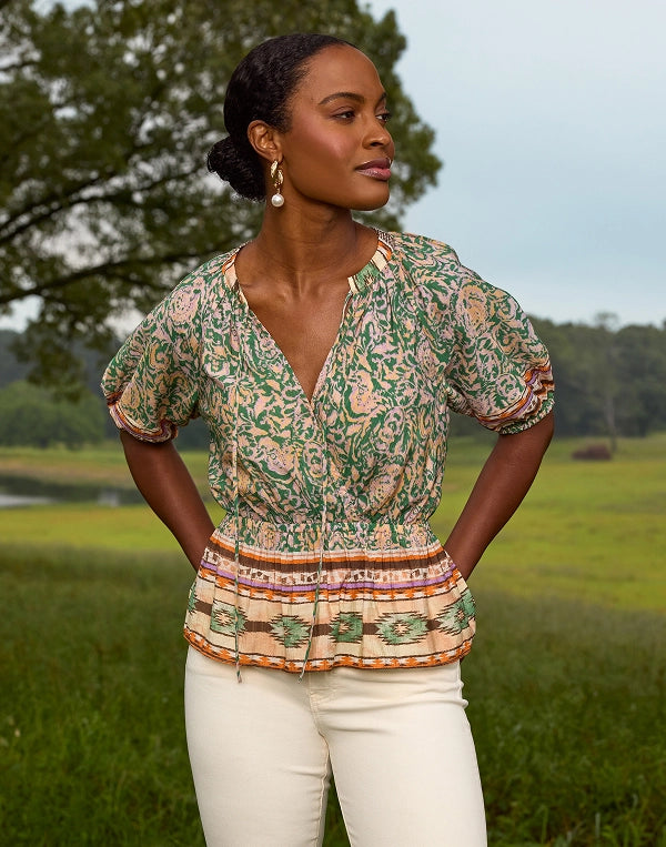 Woman wearing a patterned Robbie top and white pants standing in a field with trees in the background
