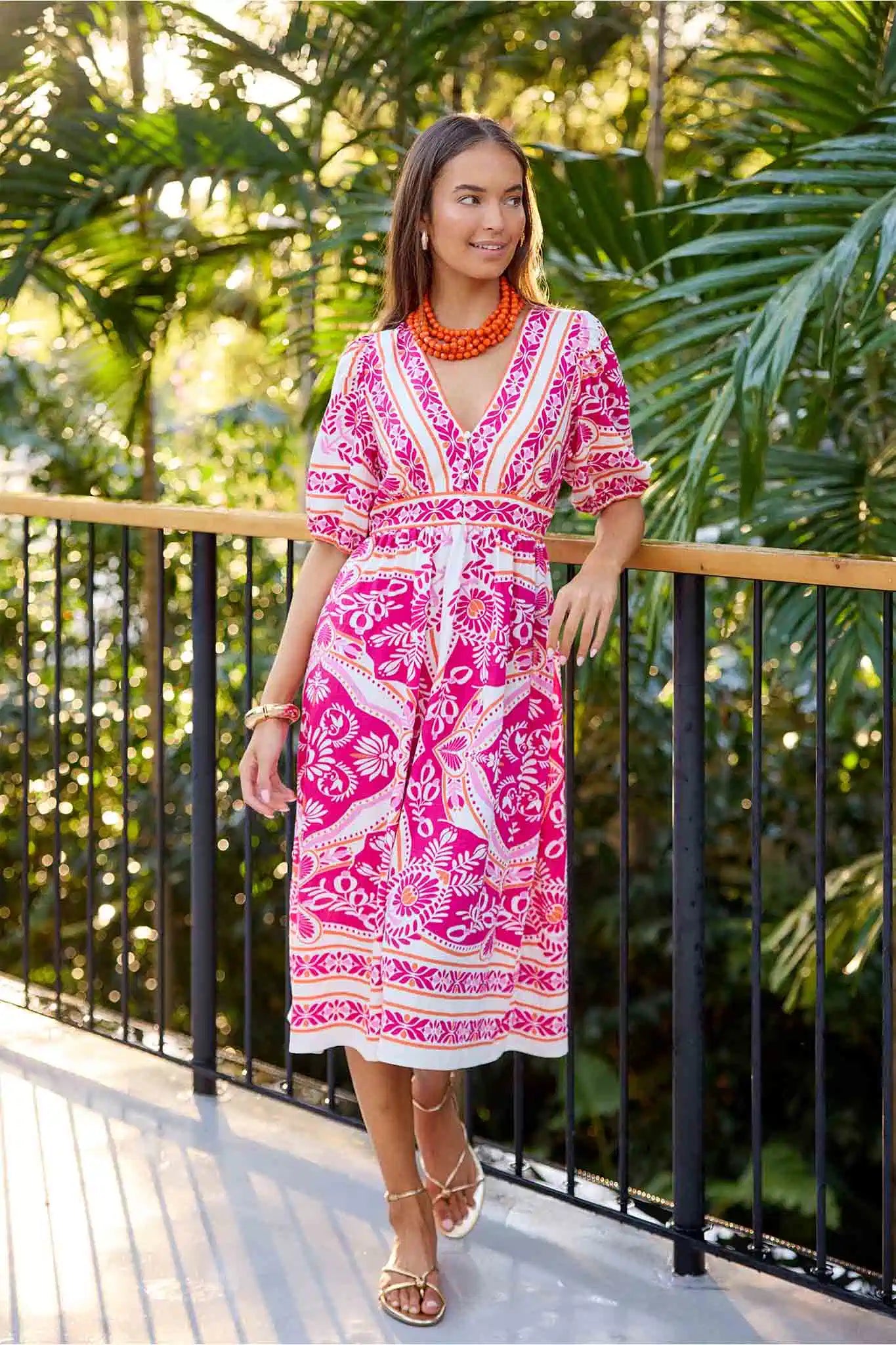 Woman in a pink and white patterned dress standing on a wooden deck with greenery in the background
