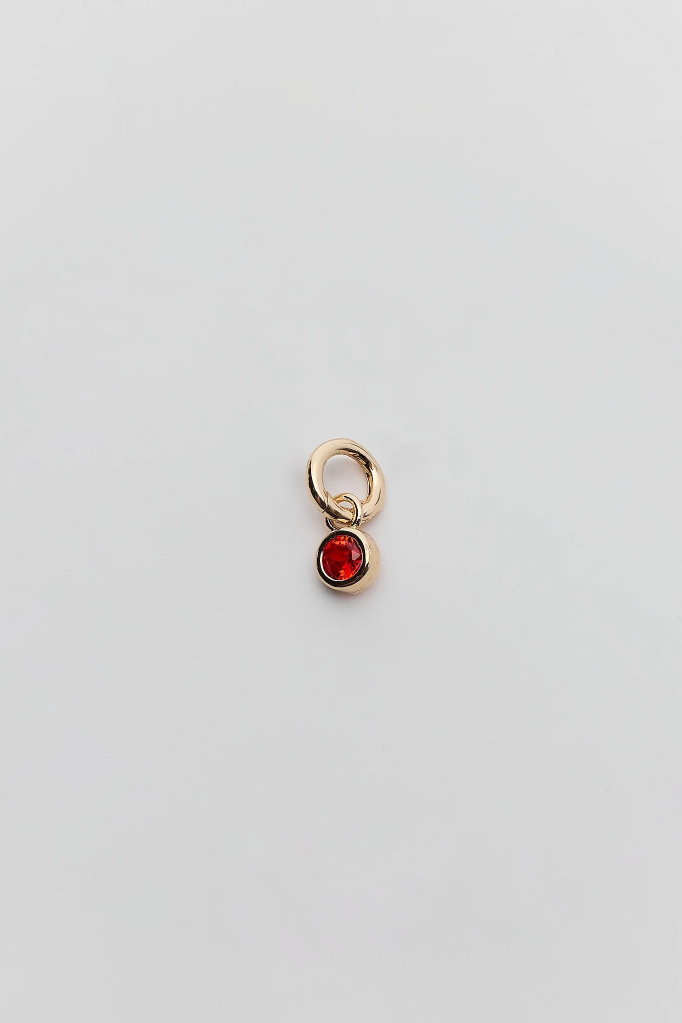 A small gold charm featuring a circular ring at the top and a dangling oval red gemstone below, set against a plain light gray background.