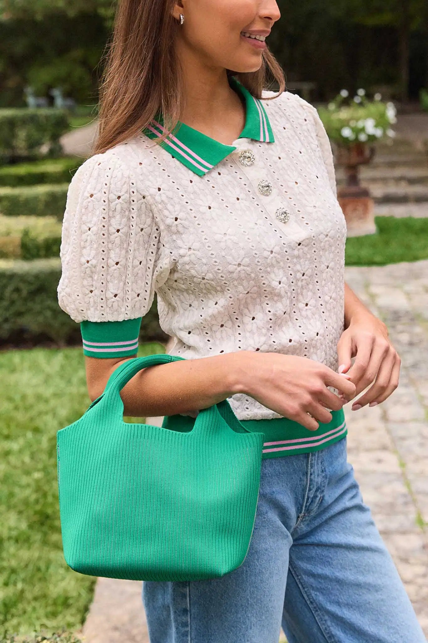 Woman holding a green handbag wearing a white blouse with green trim and blue jeans outdoors.