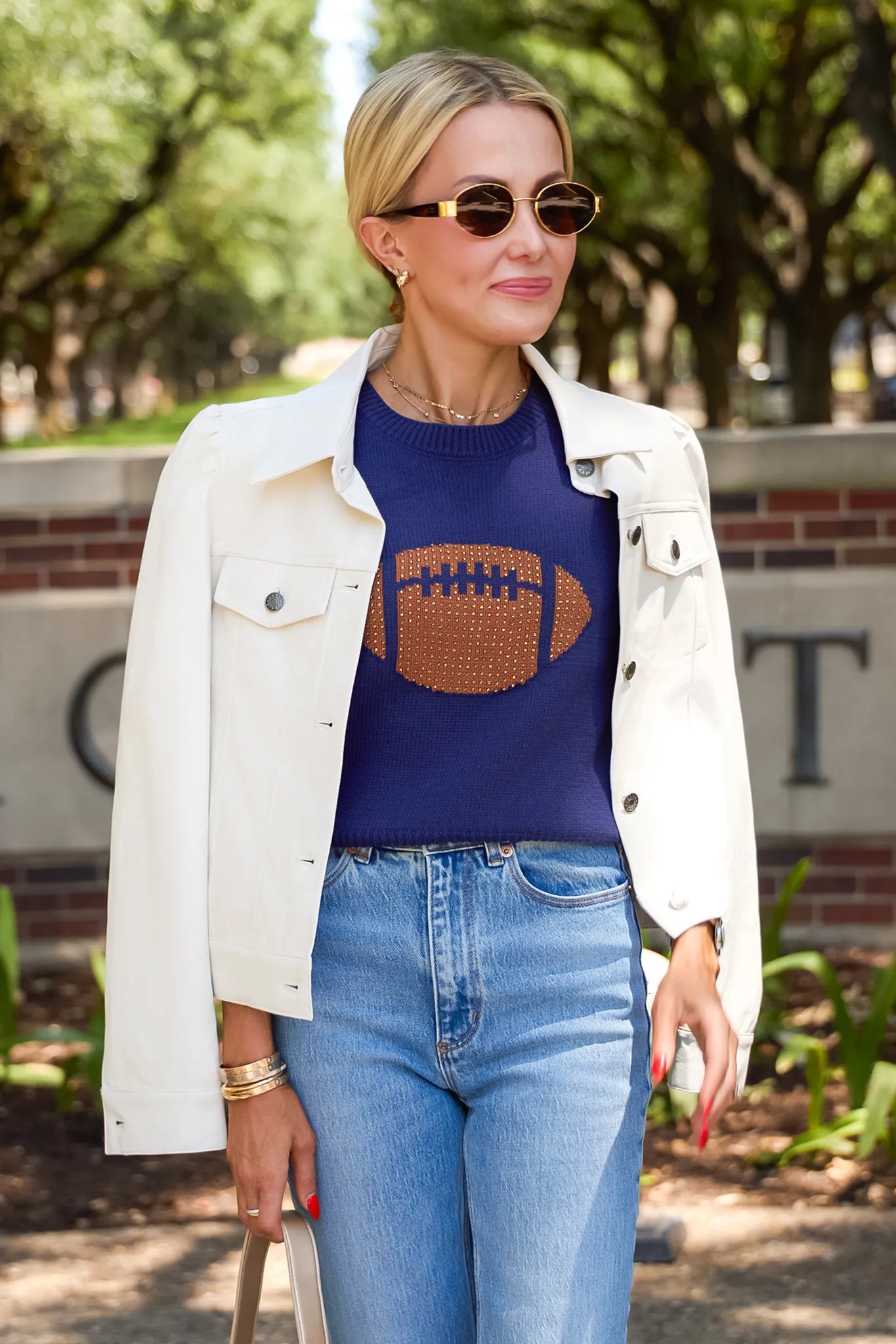 A woman wearing a navy blue crewneck sweater with a brown football graphic, styled with an off-white denim jacket and light-wash blue jeans, standing outdoors in front of a brick wall and trees.