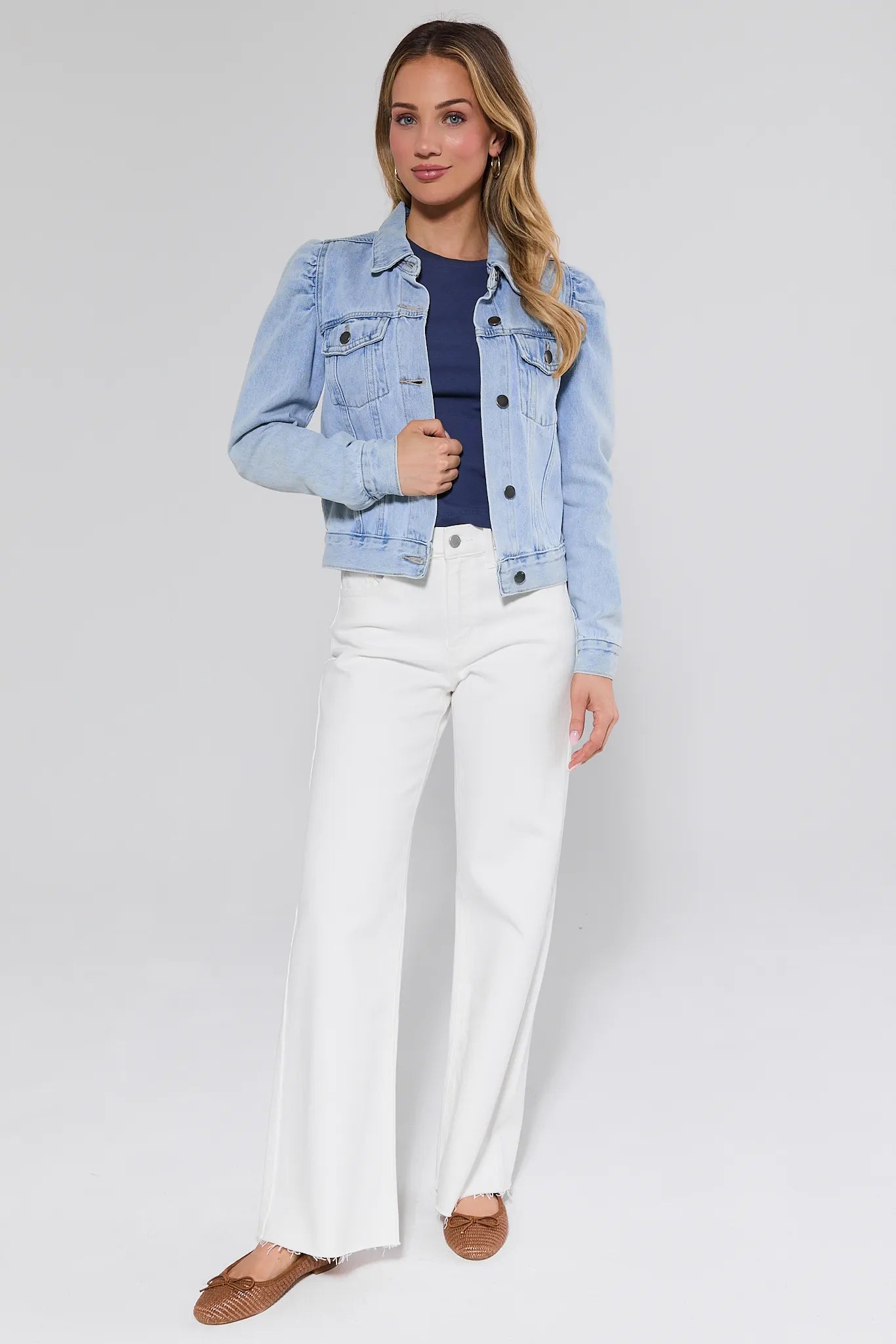 Woman wearing a light blue denim jacket, navy top, white pants, and brown shoes on a plain background