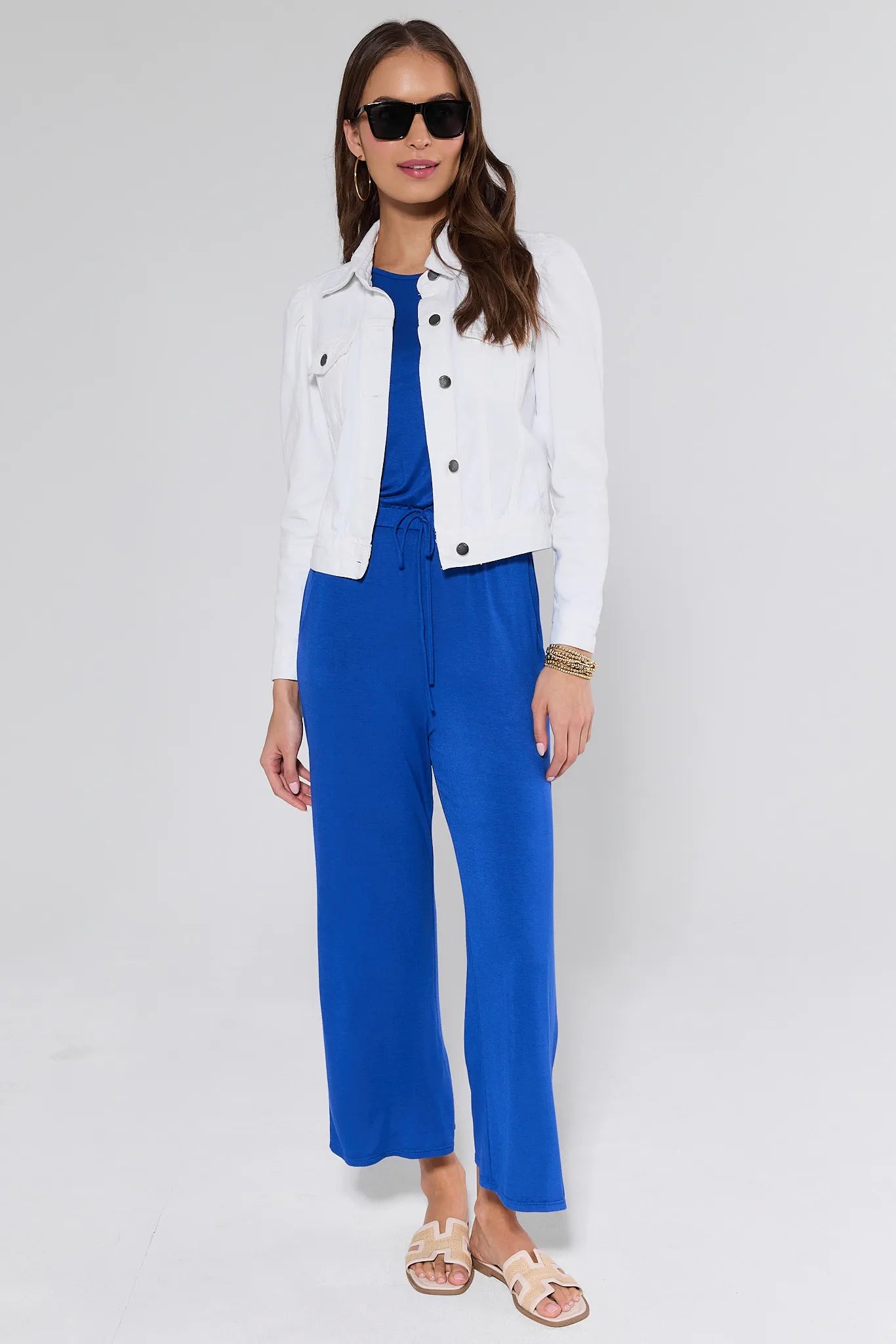Woman wearing a white jacket, blue jumpsuit, and sunglasses on a light gray background
