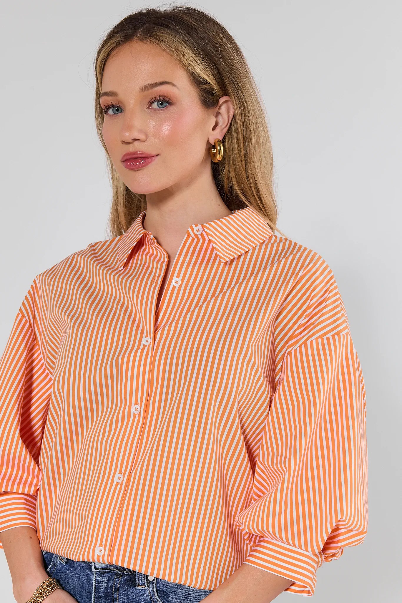 Susan Orange Striped Puff Sleeve Top: Loose-fit blouse with orange and white stripes, button-front, three-quarter gathered sleeves, and classic collar. Paired with blue jeans and gold hoop earring.