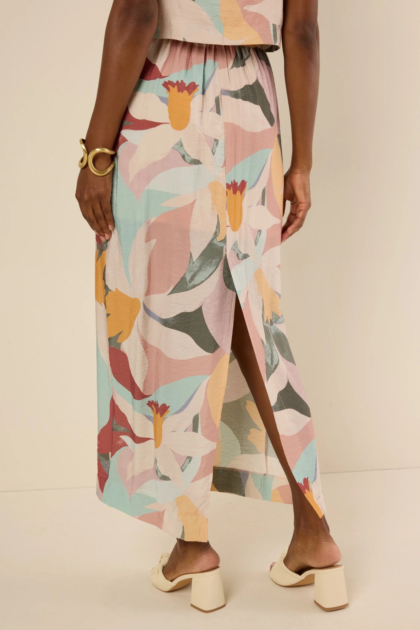 Woman modeling the Elliana Skirt, a pastel abstract floral maxi with a high side slit, paired with block-heel sandals and a gold cuff bracelet.