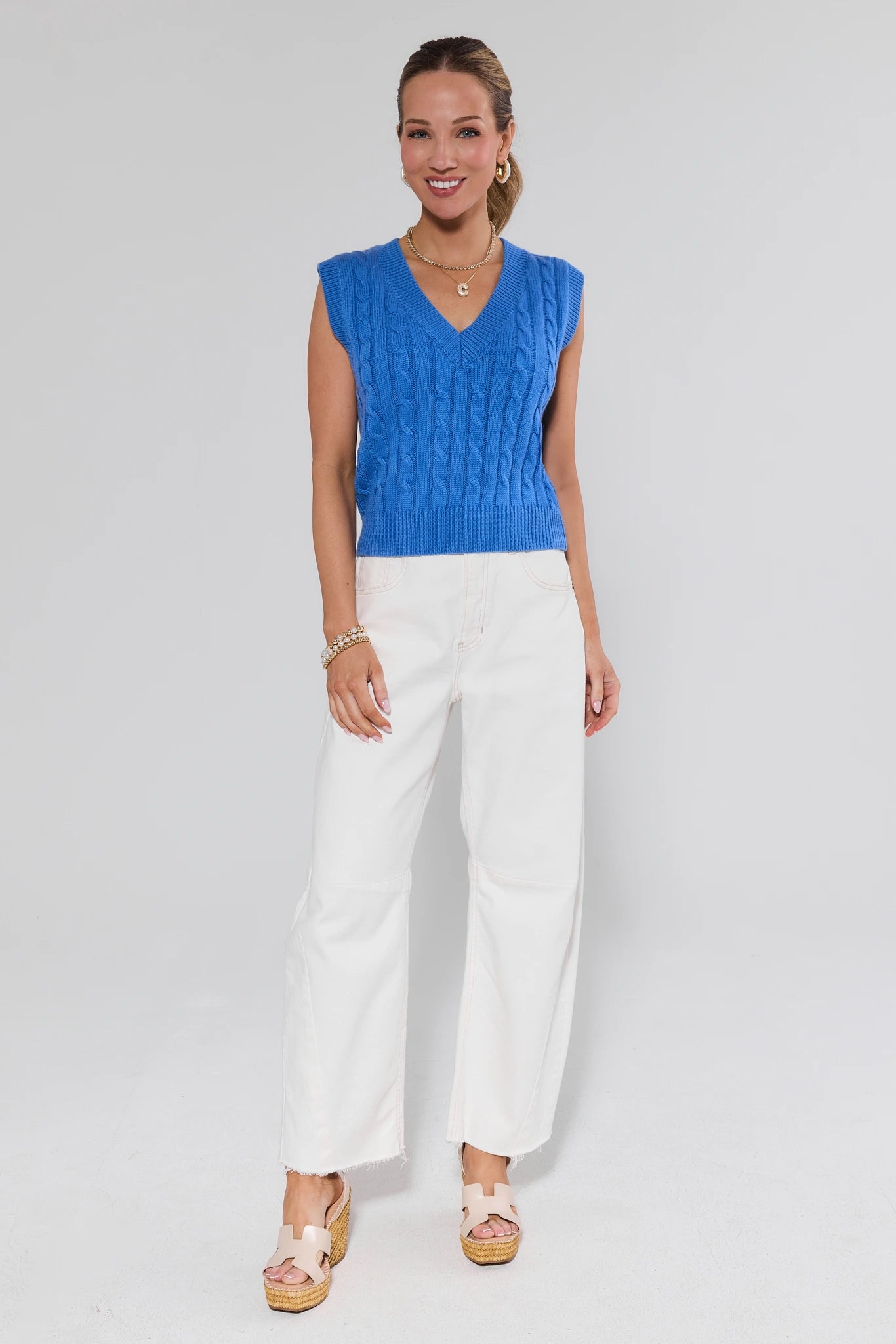 Woman wearing a blue sleeveless top and white pants on a light gray background