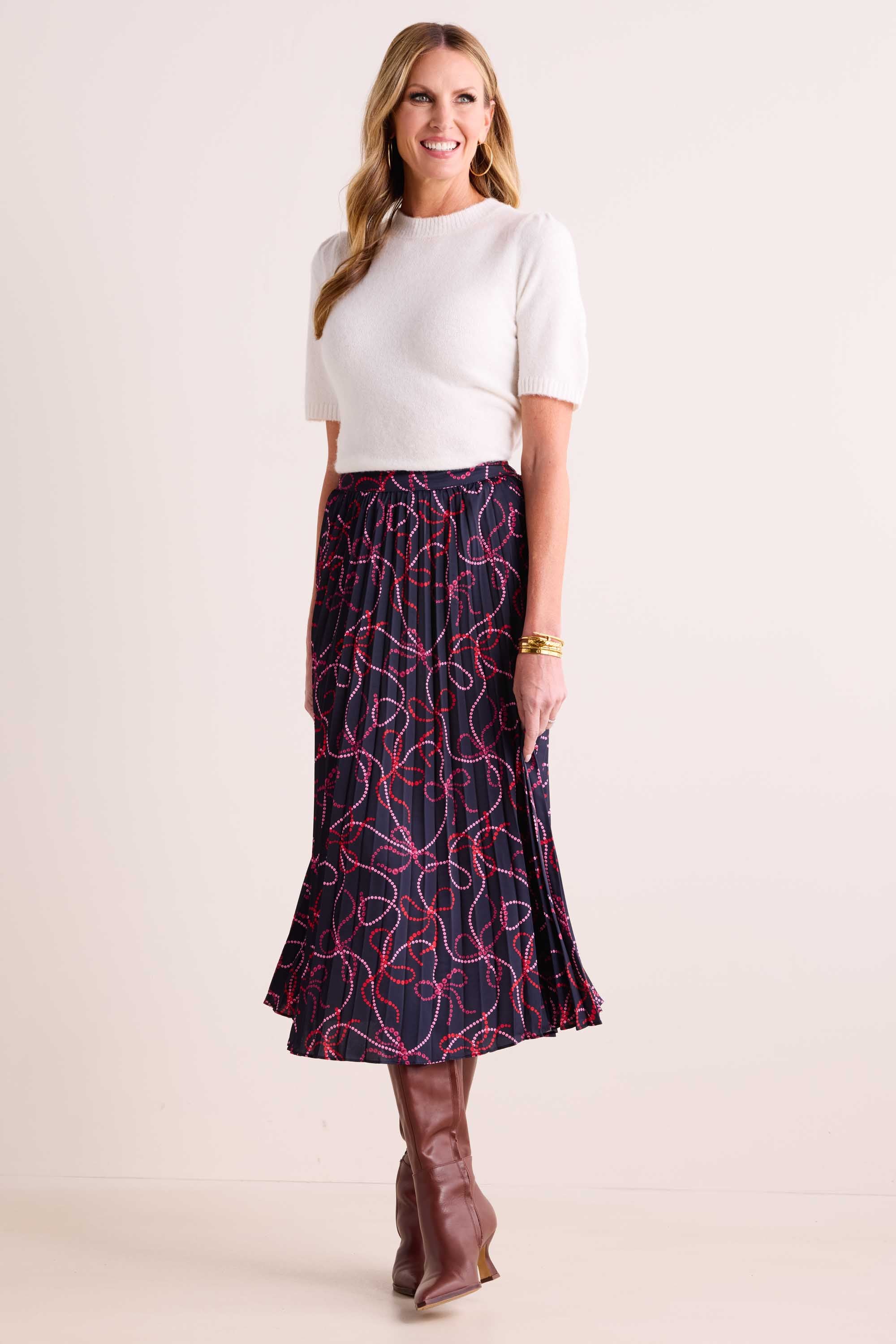 Woman wearing Ryann Skirt with pleated finish and bow print, paired with a white sweater and knee-high brown boots, accessorized with gold jewelry.