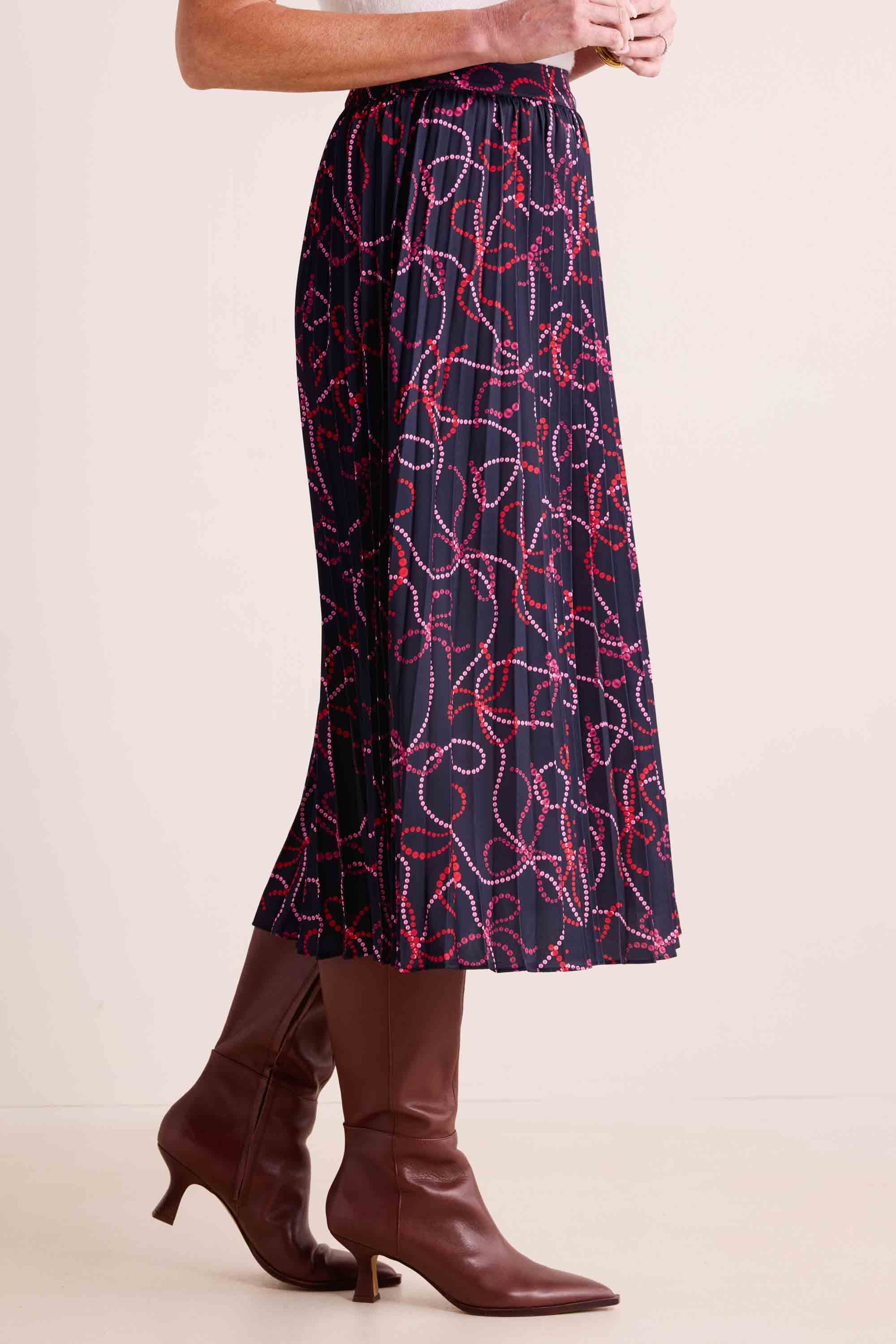 Model wearing the Ryann Skirt: a navy-blue pleated midi with red and pink bead-like lines, paired with brown pointed-toe knee-high boots.