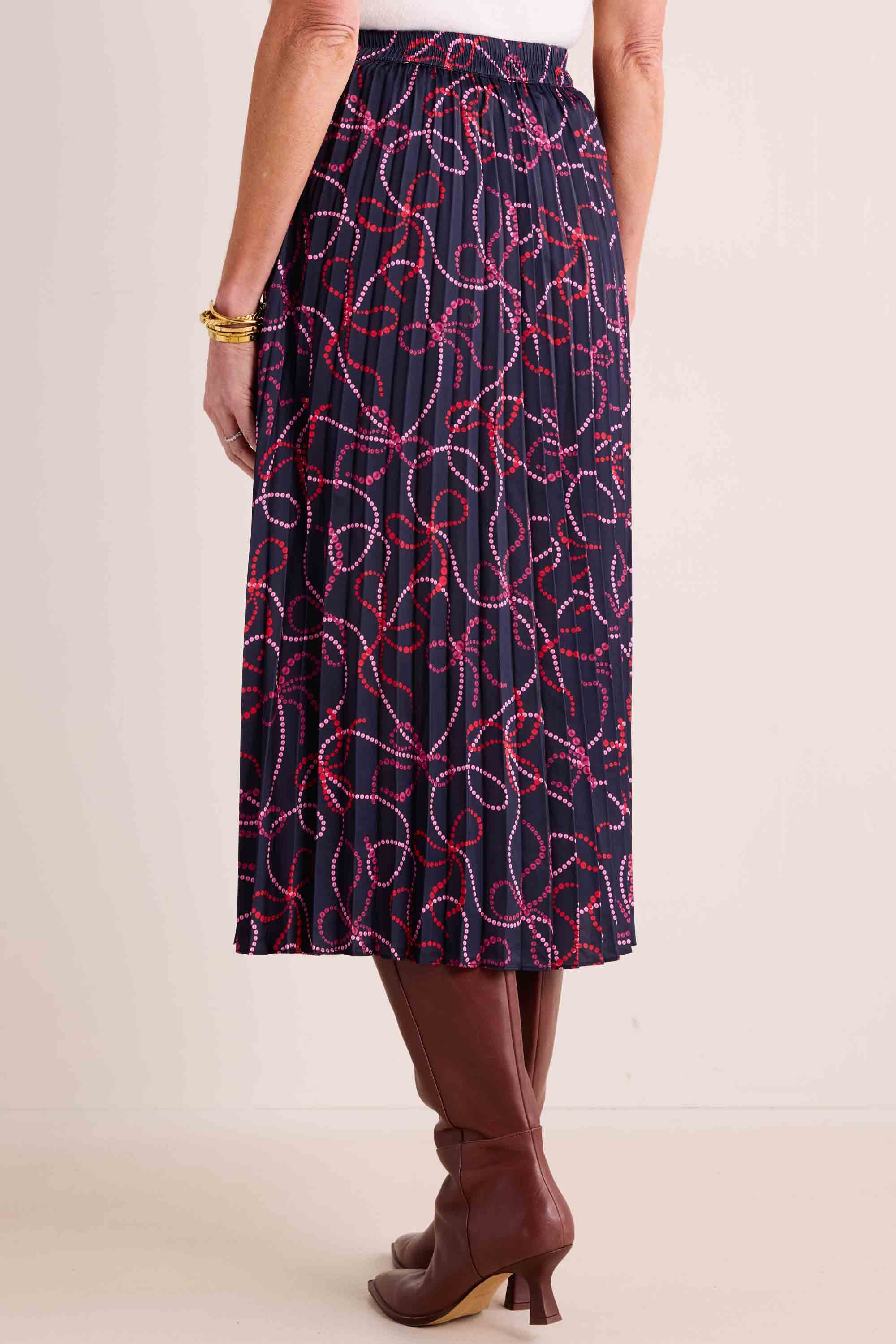 Woman wearing the Ryann Skirt with pleated design and bow print, paired with brown knee-high boots, showcasing a chic, versatile fashion ensemble.