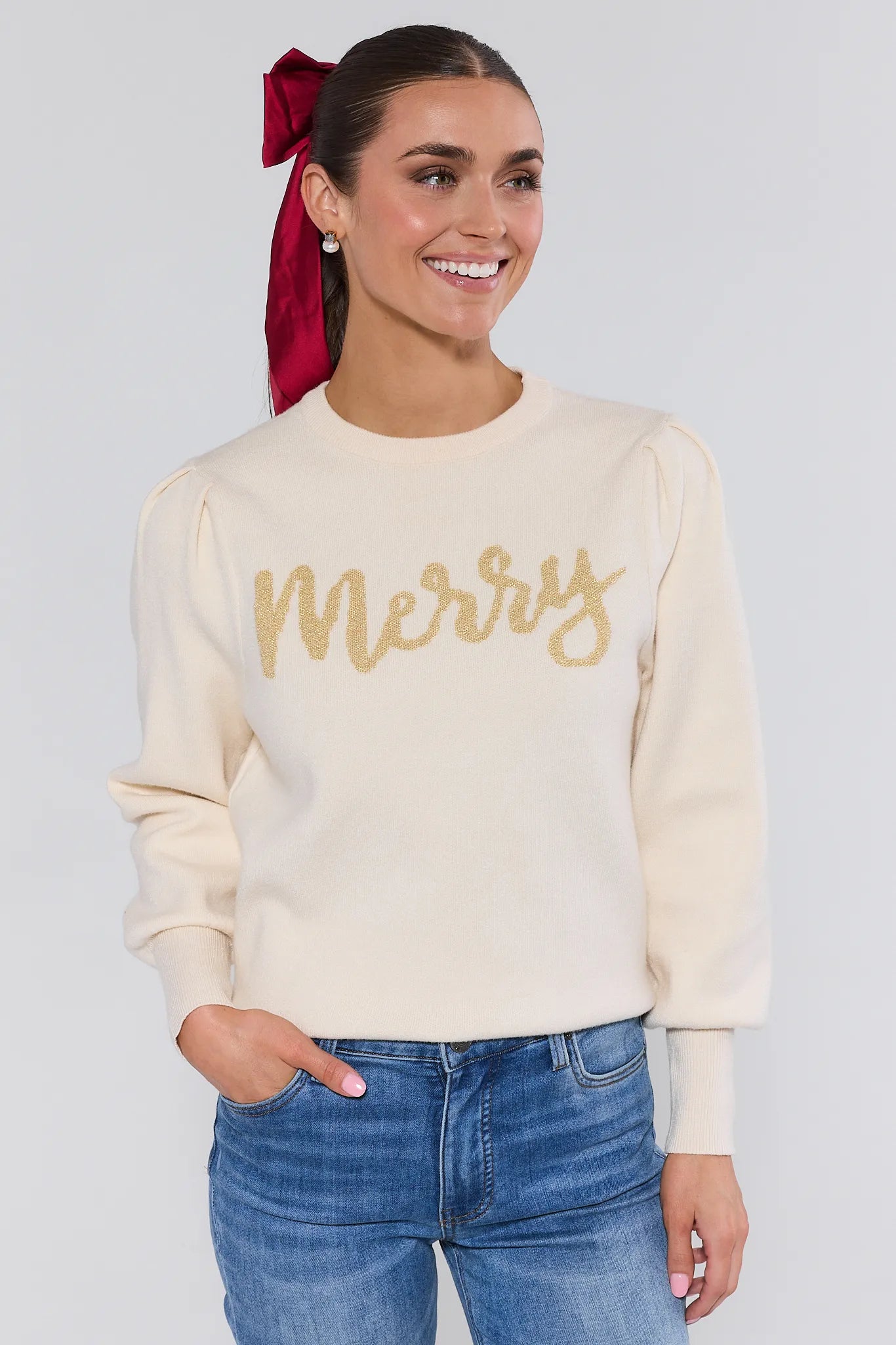 Woman wearing a cream sweater with 'Merry' text, blue jeans, and a red bow on a plain background