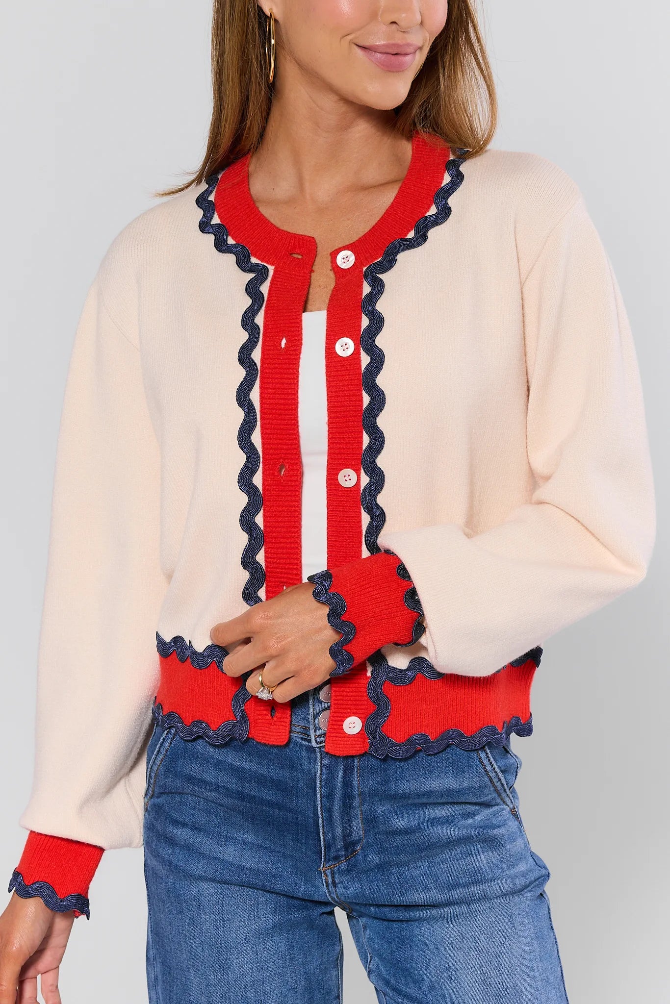 Woman wearing a cream cardigan with red and navy trim, white shirt, and blue jeans on a light gray background.