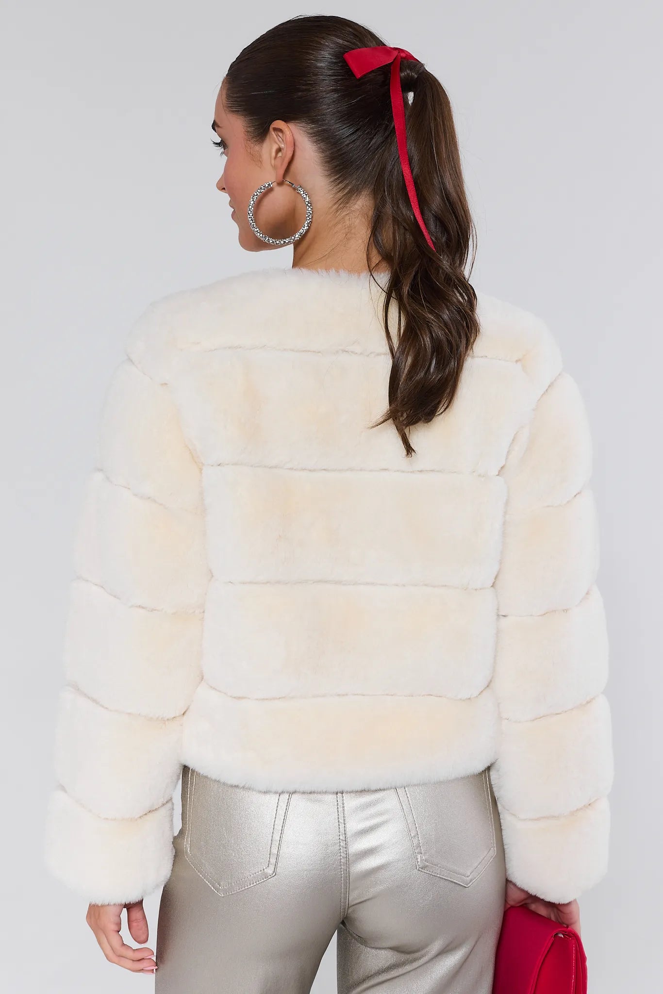 Woman wearing a cream fur jacket and silver pants with a red clutch against a white background