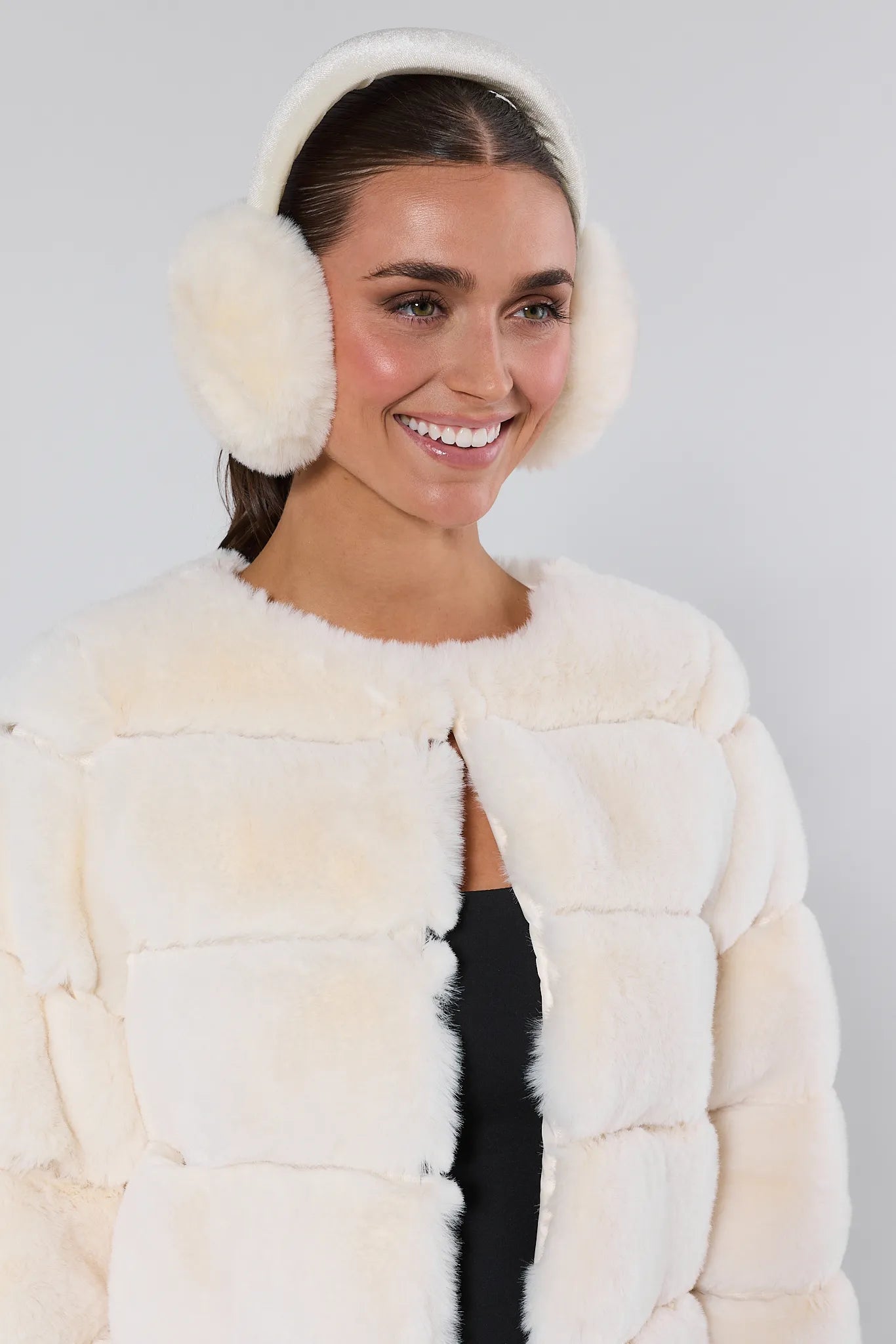 Woman wearing a cream fur coat and white earmuffs against a gray background