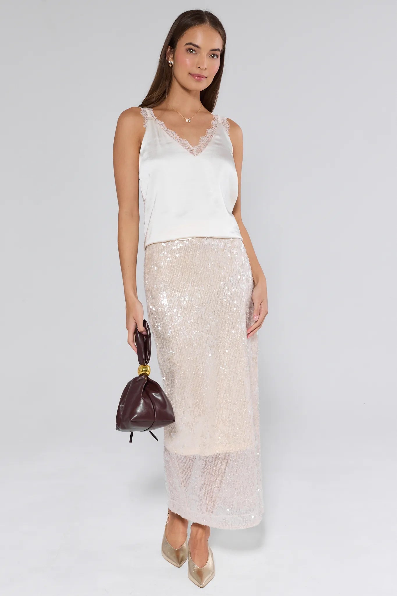 Woman wearing a white sleeveless top and beige sequin skirt holding a brown handbag on a white background
