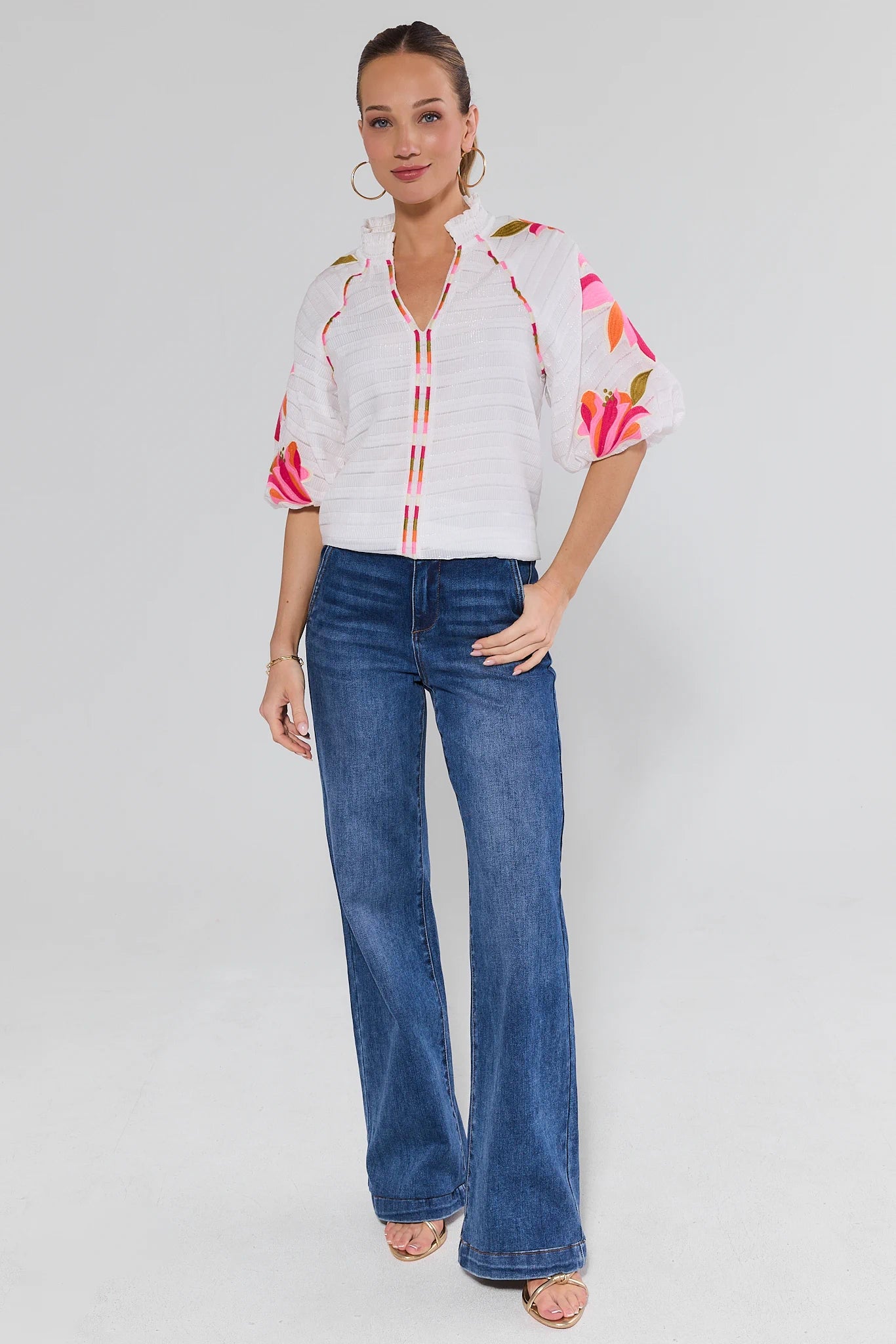Woman wearing a white blouse with floral sleeves and blue jeans on a gray background