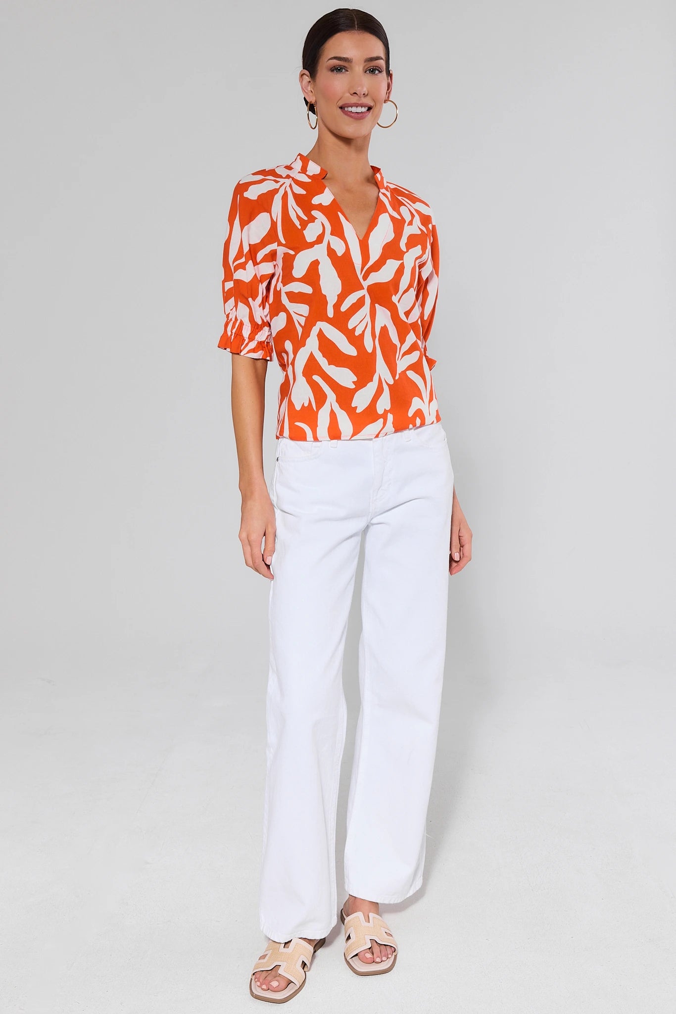 Woman wearing an orange and white patterned trish blouse with white pants on a gray background