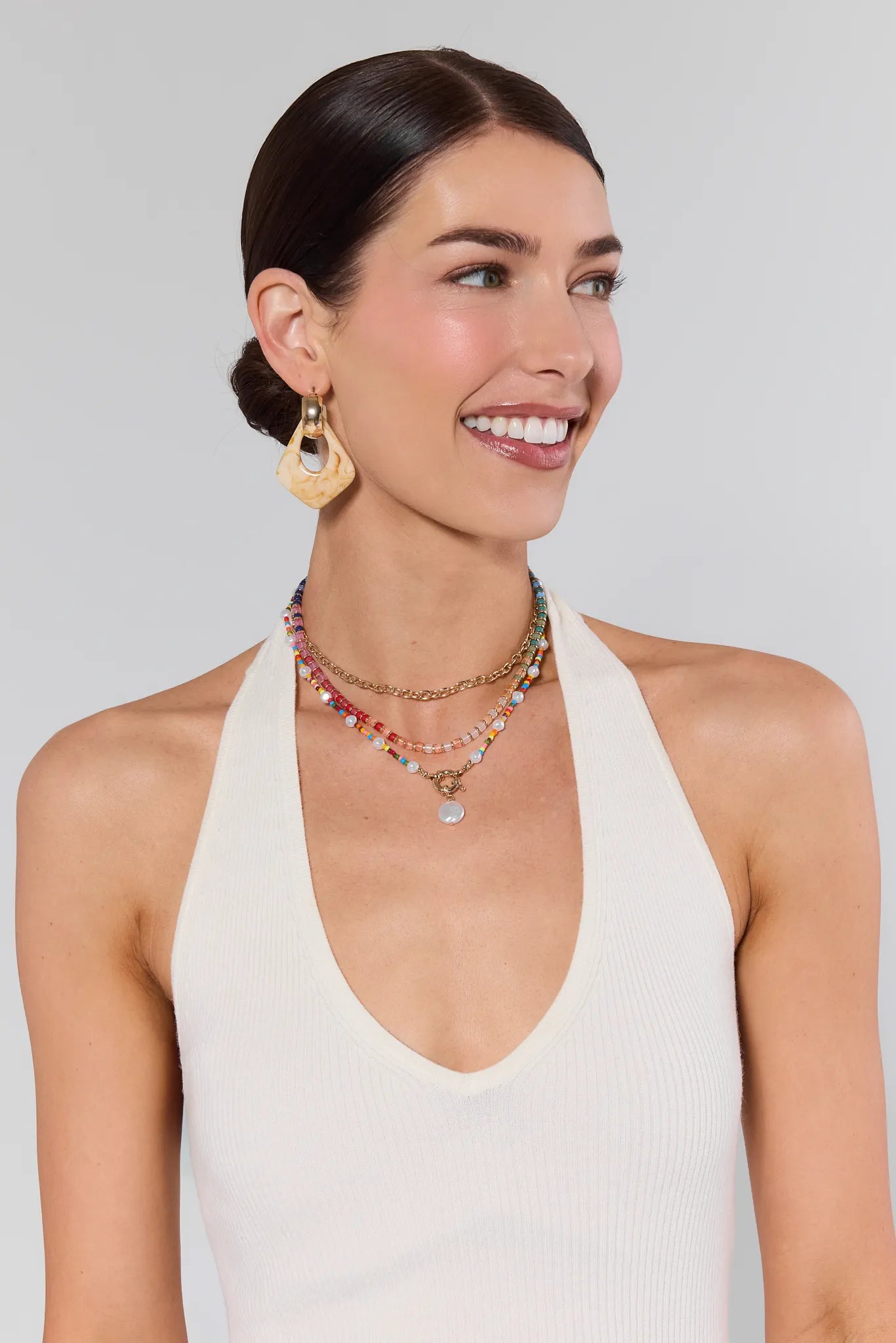 Woman wearing gold earrings and multiple necklaces against a plain background