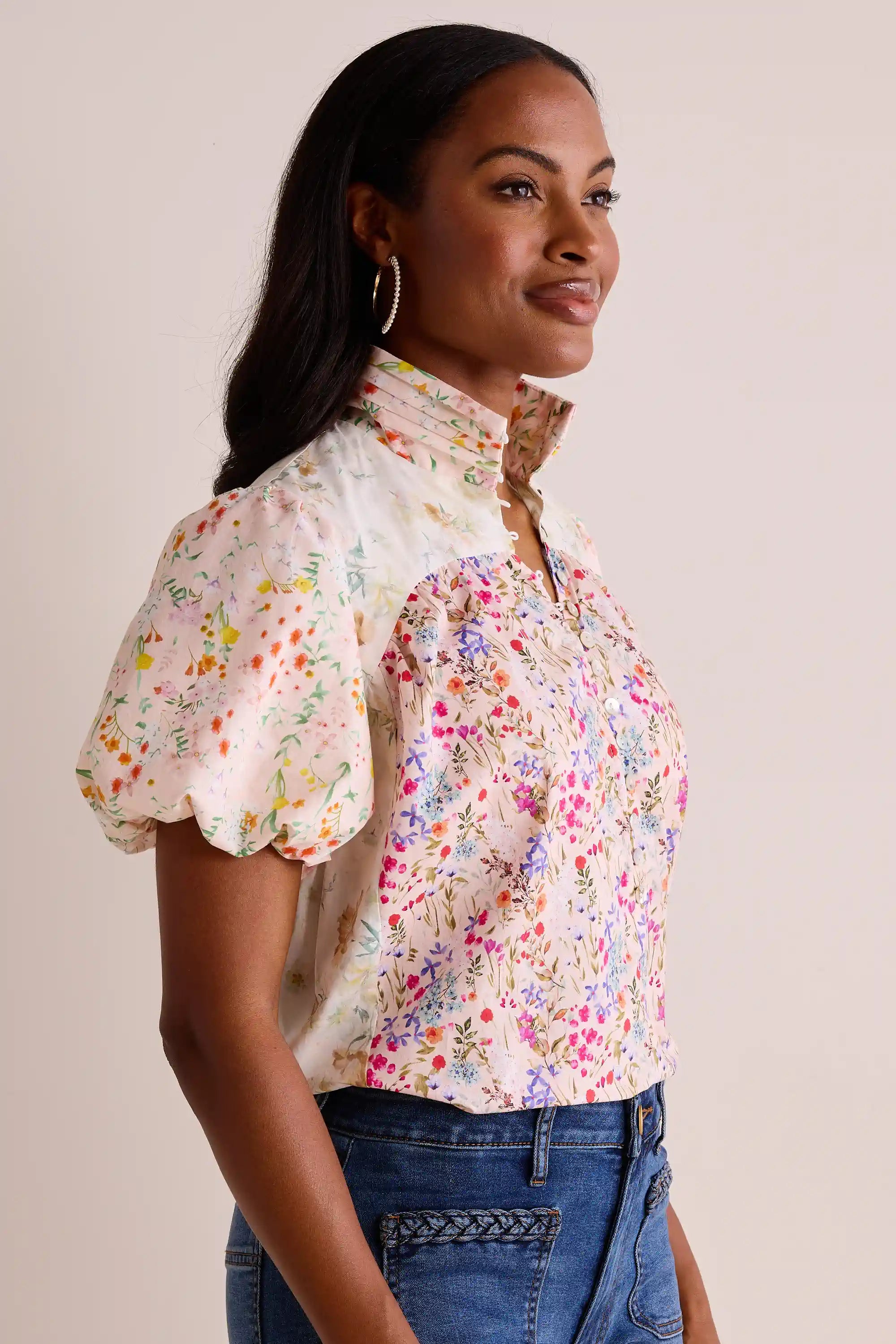 Model wearing the Margie Floral Print Puff Sleeve Top with multicolored wildflowers, stand-up collar, and button-front, paired with medium-wash jeans and hoop earrings.
