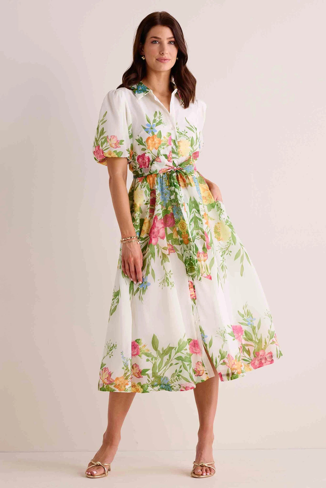 Woman wears Claudette Dress, a white midi shirt dress with colorful florals, puff sleeves, collar, and tie waist, accessorized with gold sandals and jewelry.