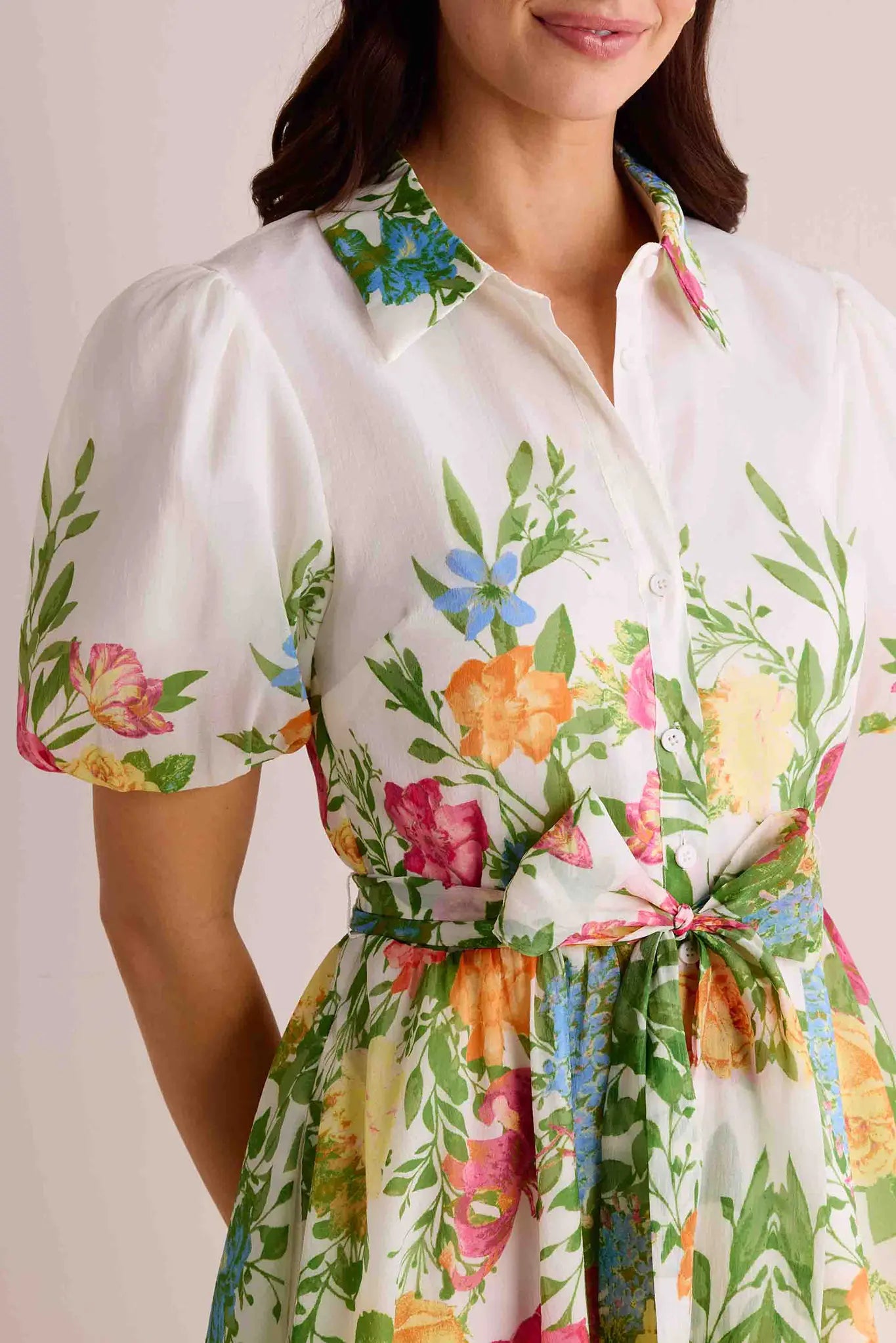 Woman in Claudette Dress: a white, short-sleeved, button-down shirt-dress with a floral pattern, puffed sleeves, and a fabric belt tied at the waist.