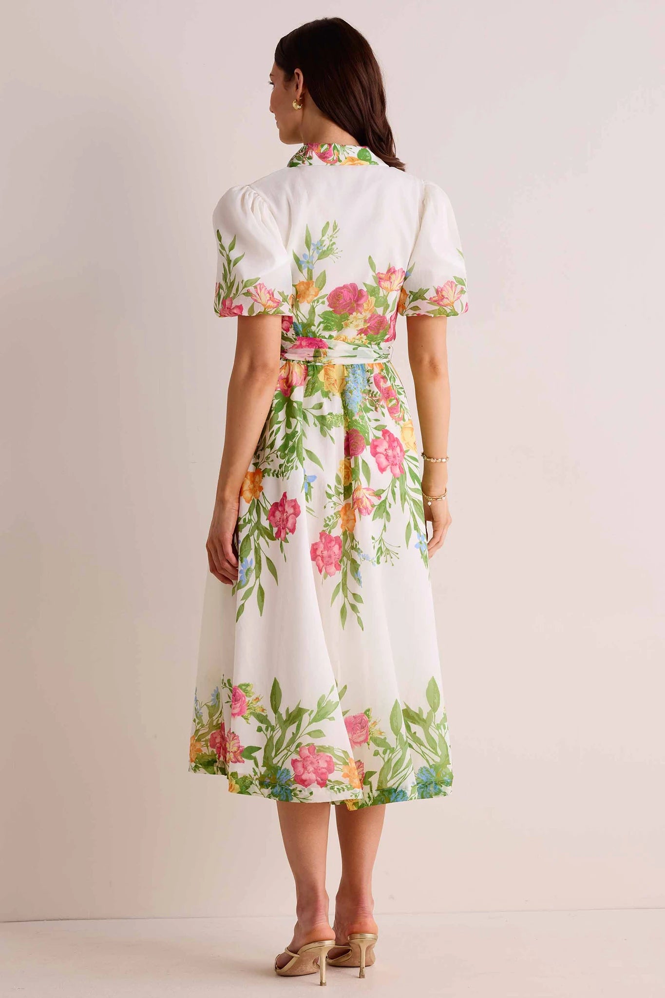 Woman wearing Claudette Dress, a white shirt-dress with floral print, puff sleeves, self-tie belt, accessorized with gold earrings and sandals, viewed from behind.