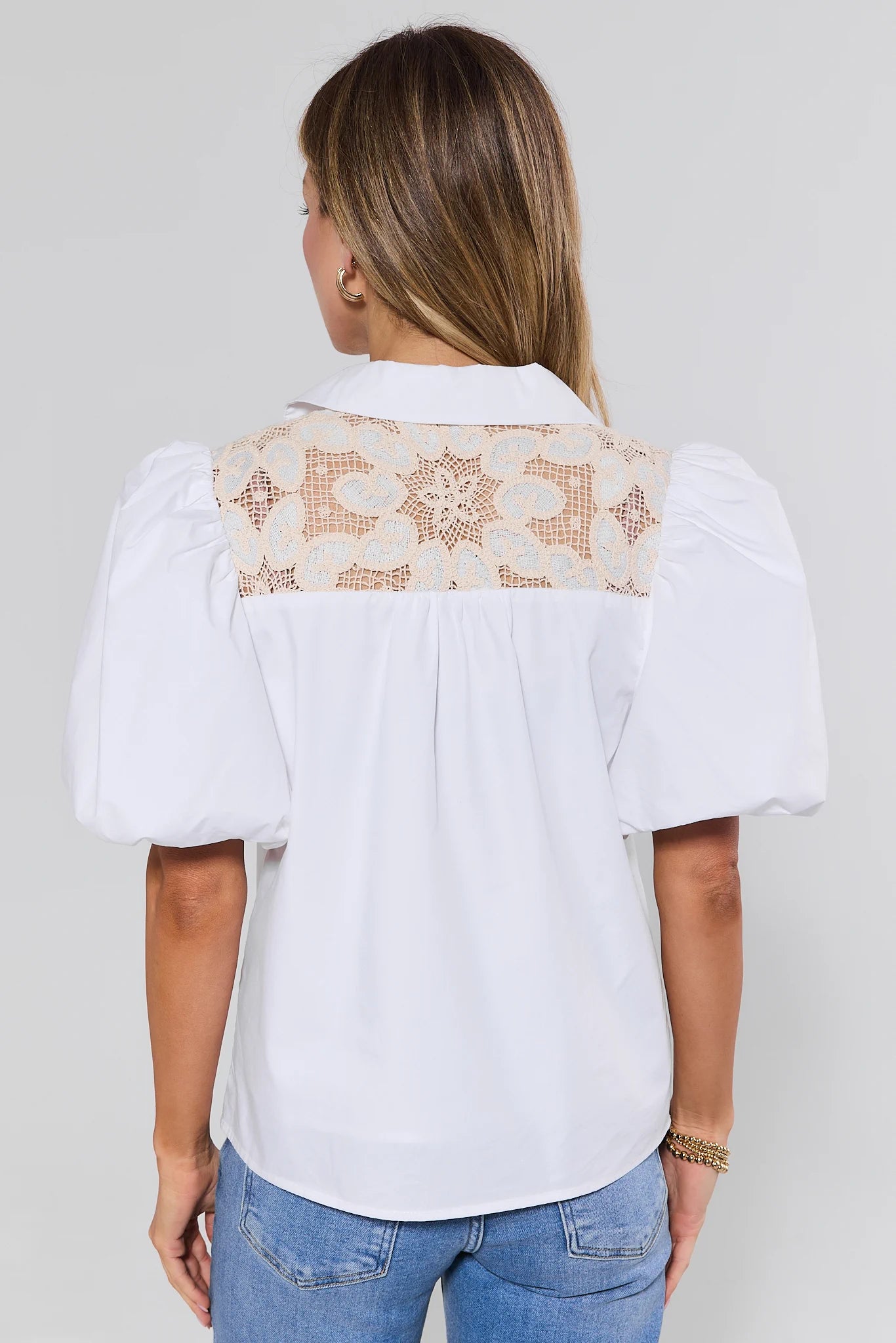 Woman wearing a white blouse with lace detailing on the back, standing against a plain background.