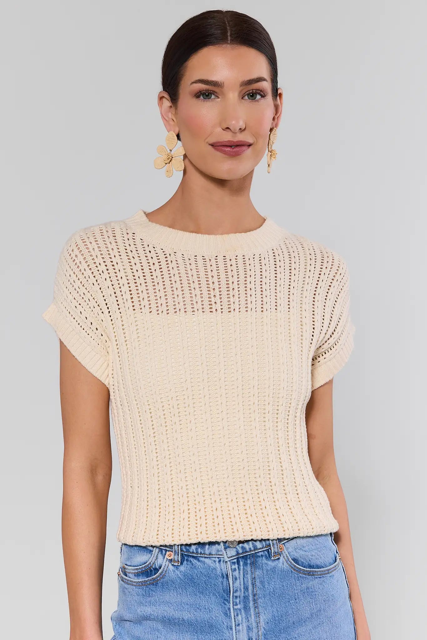 Woman wearing a beige knitted beatrice top and blue jeans against a plain background